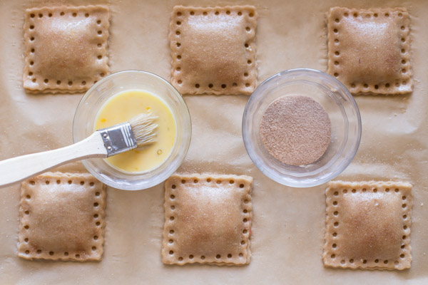 Whole Wheat Strawberry Rhubarb Fruit Pockets on a parchment paper lined baking sheet, with a bowl of egg wash with a brush in it and a bowl of cinnamon sugar.