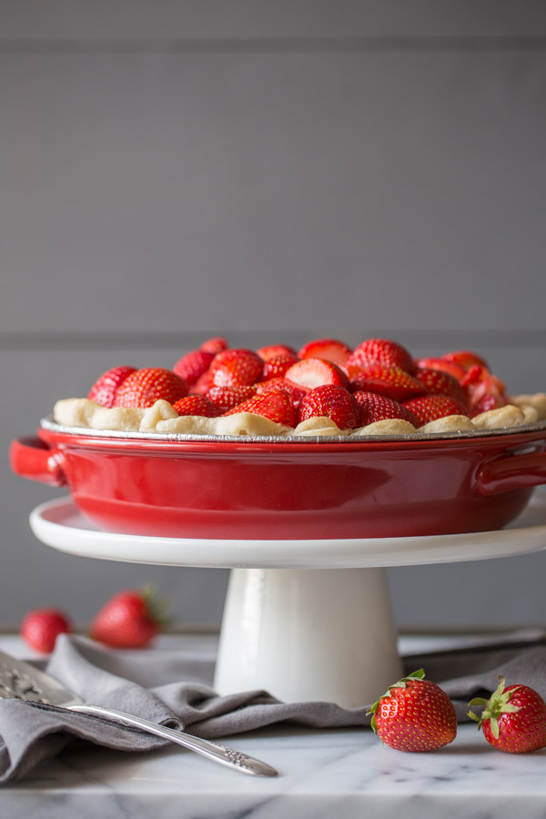 A Fresh Strawberry Pie in a pie plate, sitting on top of a cake stand, with a serving utensil and whole strawberries next to it.  