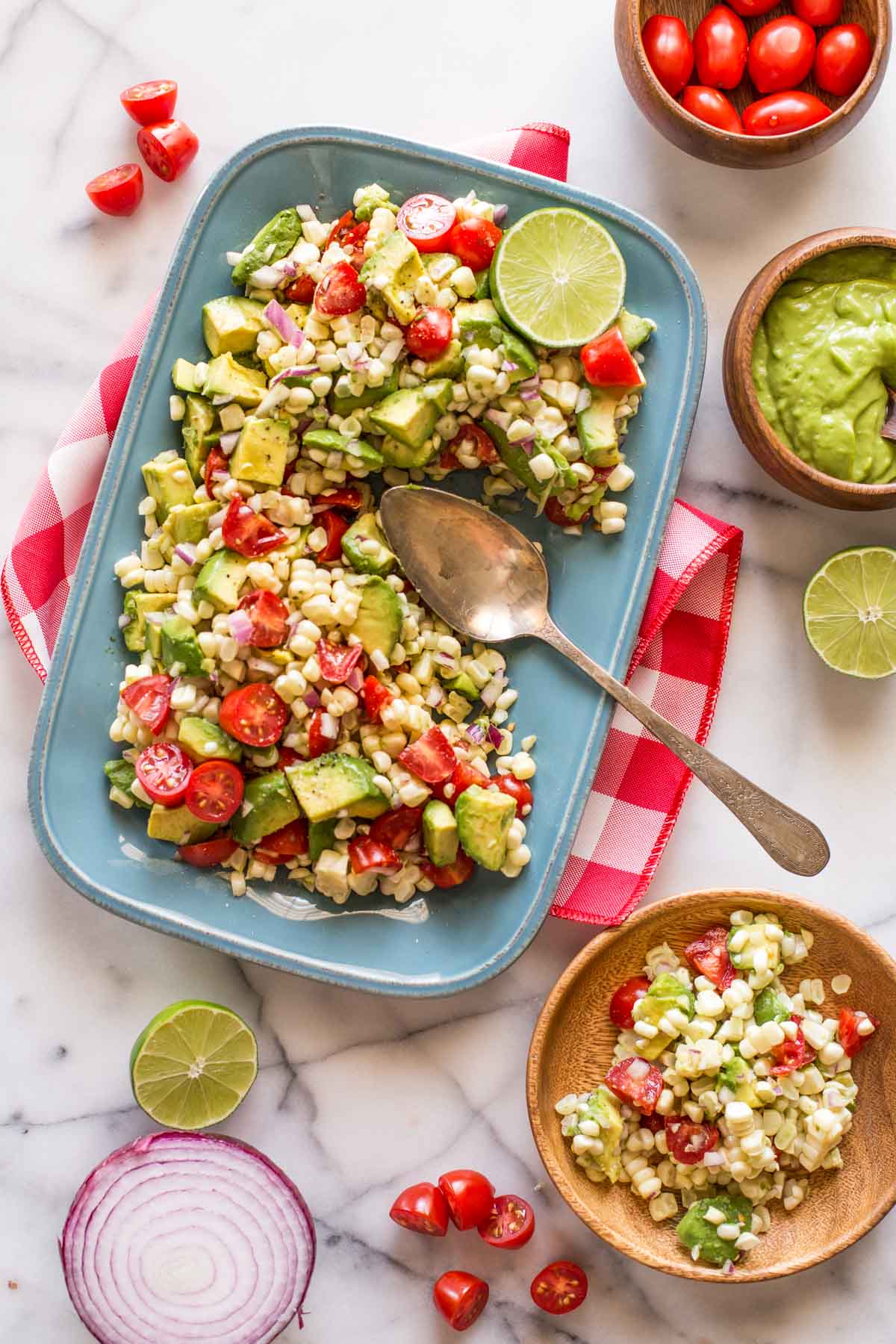 Corn, Tomato, and Avocado Salad on a platter with a serving spoon, sitting next to a small bowl of grape tomatoes, a small bowl of the avocado dressing, and a wood plate with a serving of the salad on it.