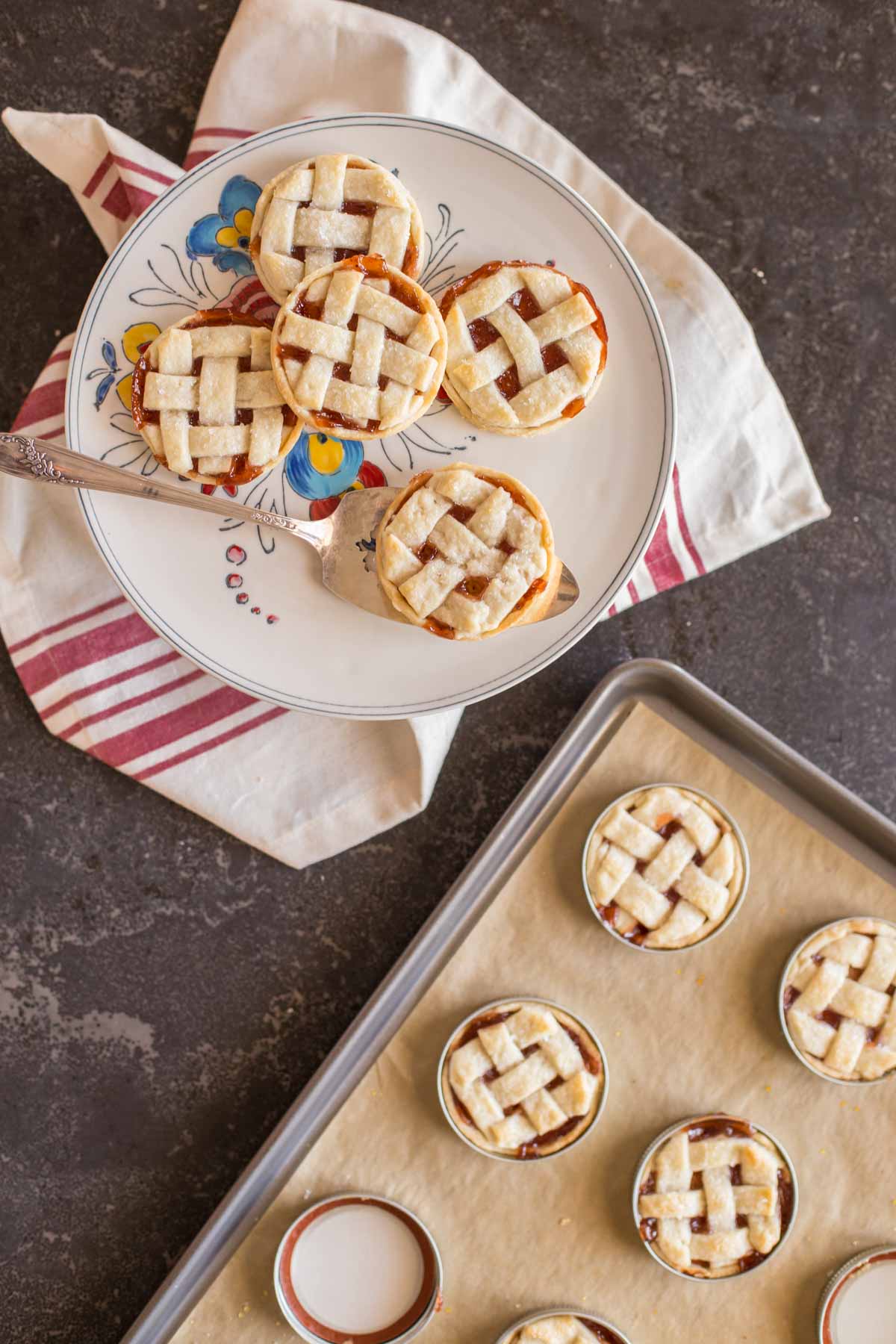 Mini Mason Jar Strawberry Pies on a plate with a serving utensil, sitting next to a baking sheet of more Mini Mason Jar Strawberry Pies.
