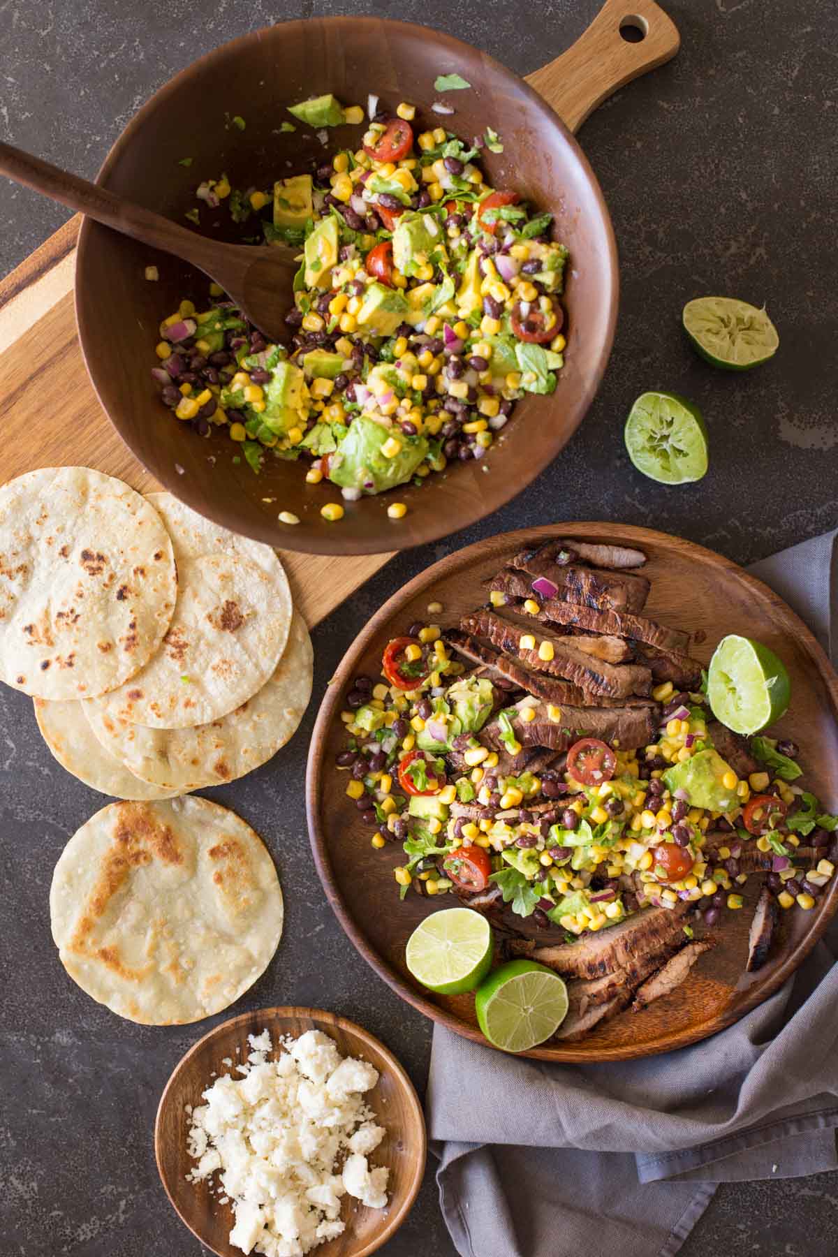 A large wood plate of sliced grilled marinated flank steak and avocado salad with limes cut in half, sitting next to a large wood bowl of avocado salad, some small taco tortillas and a small wood plate of crumbled queso fresco. 