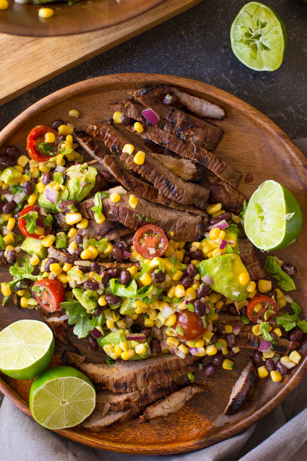 A large wood plate of sliced grilled marinated flank steak and avocado salad with limes cut in half. 