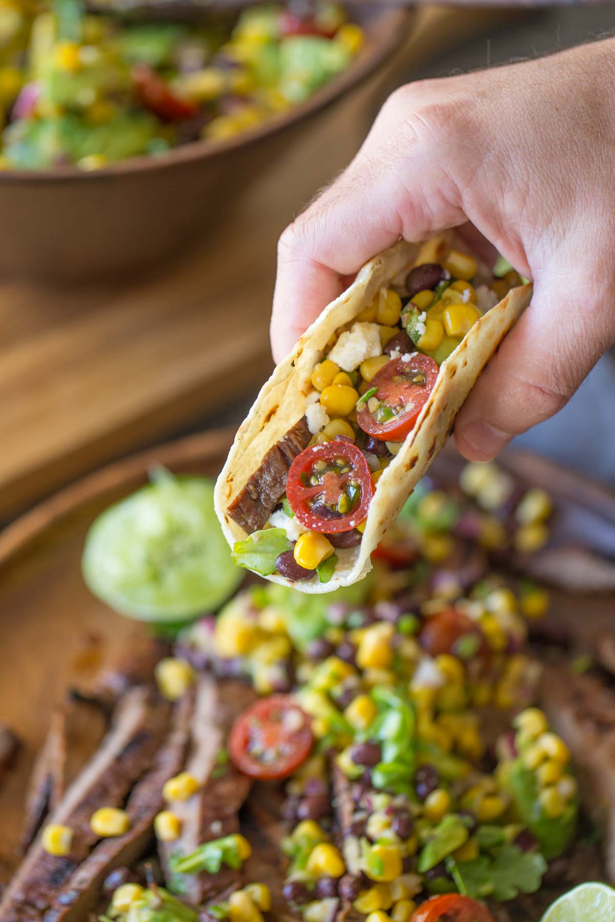 A Flank Steak Taco being held over a large wood plate of sliced grilled marinated flank steak and avocado salad.