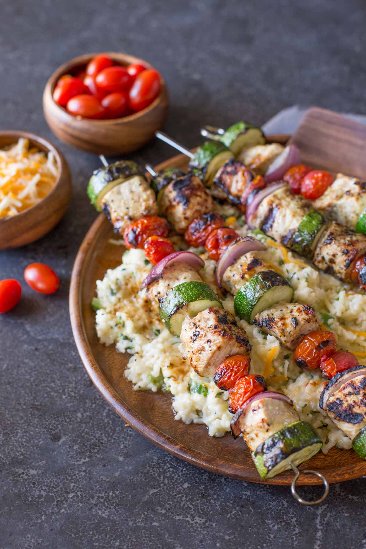 Pork Tenderloin Kabobs served over Cheesy Broccoli Rice on a large wood plate, with a small wood bowl of shredded cheese and a small wood bowl of grape tomatoes in the background.