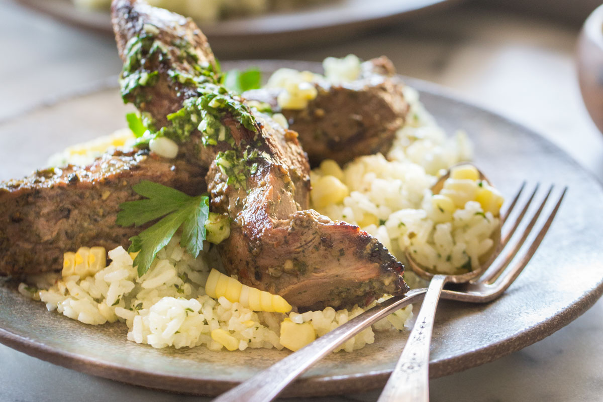 Brazilian Grilled Flank Steak resting on a bed of Chimichurri Rice, on a plate with a fork and a spoon.