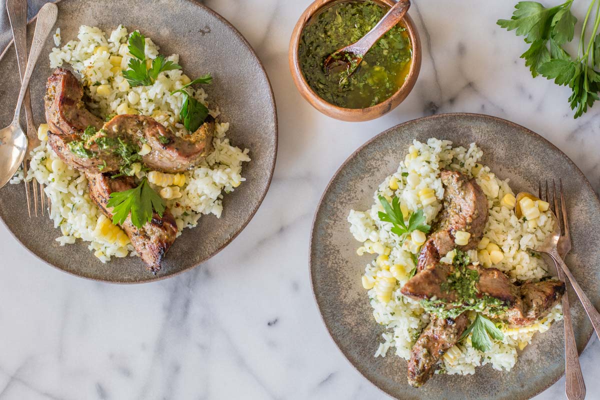 Two plates of Brazilian Grilled Flank Steak resting on a bed of Chimichurri Rice, with a fork and spoon on each plate, and a small bowl of chimichurri next to the plates.