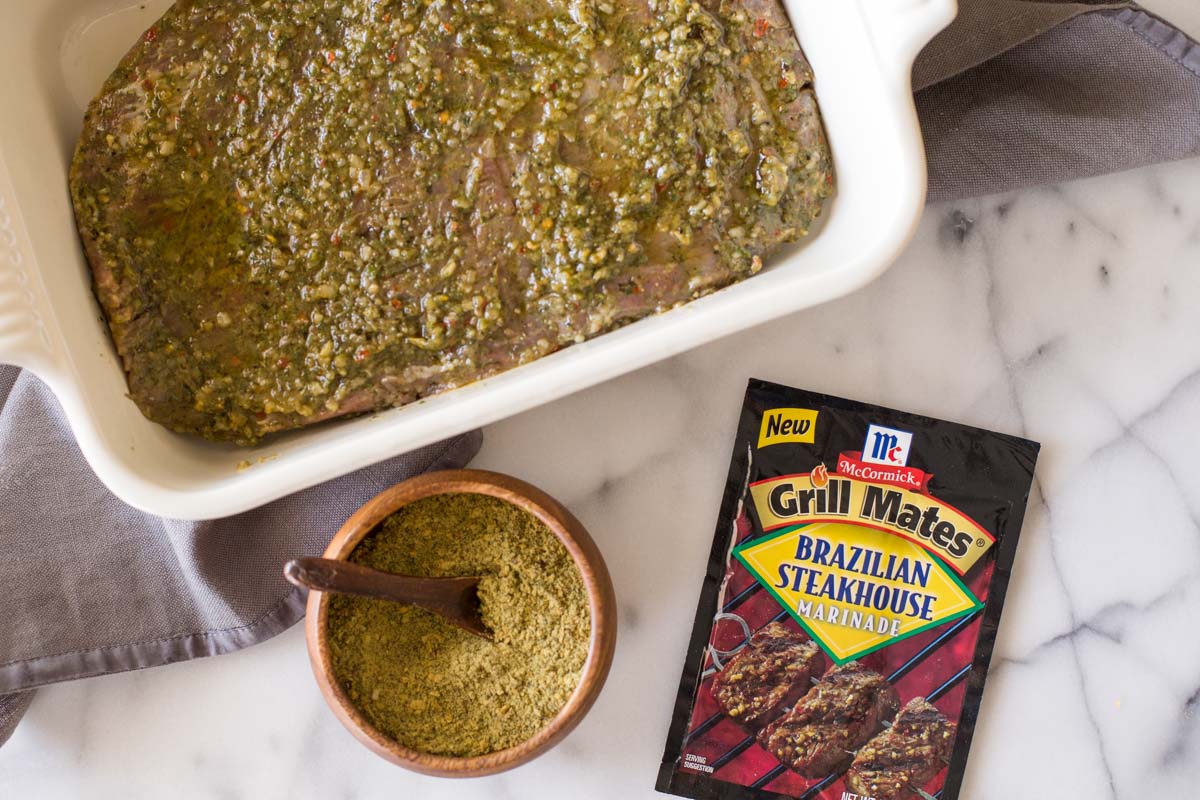 Flank steak in a baking dish, covered with the marinade, with a packet of the McCormick Brazilian Steakhouse Marinade next to the dish, as well as a small wood bowl of the spices with a wooden spoon in it.