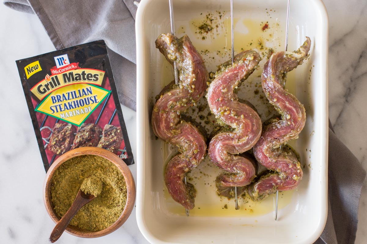 Marinated flank steak sliced into one inch strips, and threaded onto skewers in a baking dish, with a packet of the McCormick Brazilian Steakhouse Marinade next to the dish, as well as a small wood bowl of the spices with a wooden spoon in it.