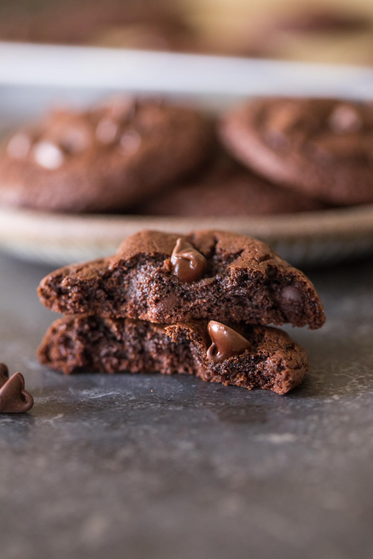 A One Bowl Triple Chocolate Brownie Cookie broken in half and stacked, with a plate of cookies in the background. 