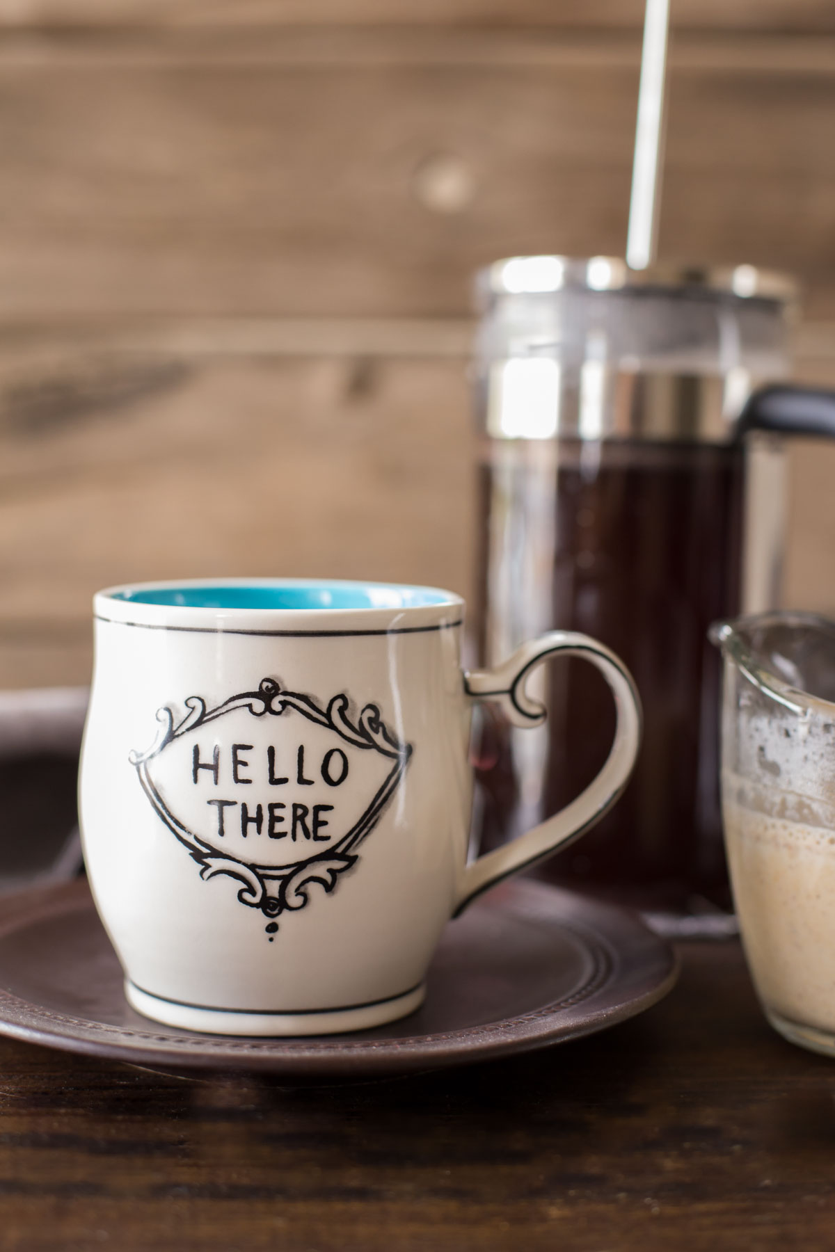 A coffee mug sitting on a plate, with a glass pour cup of Homemade Pumpkin Spice Coffee Creamer sitting next to it, and a French Press coffee maker in the background.