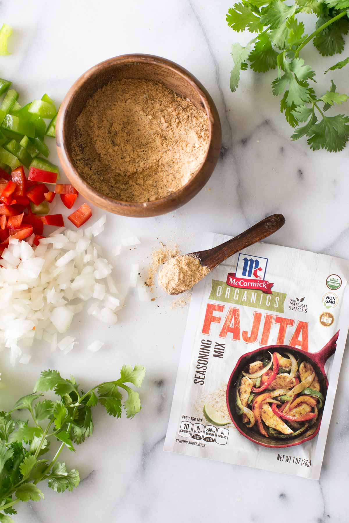 A packet of McCormick Organics Fajita Seasoning Mix, with a small wood spoon of the seasoning mix next to a small wood bowl of the seasoning mix, along with copped green and red peppers, chopped onions and fresh cilantro.