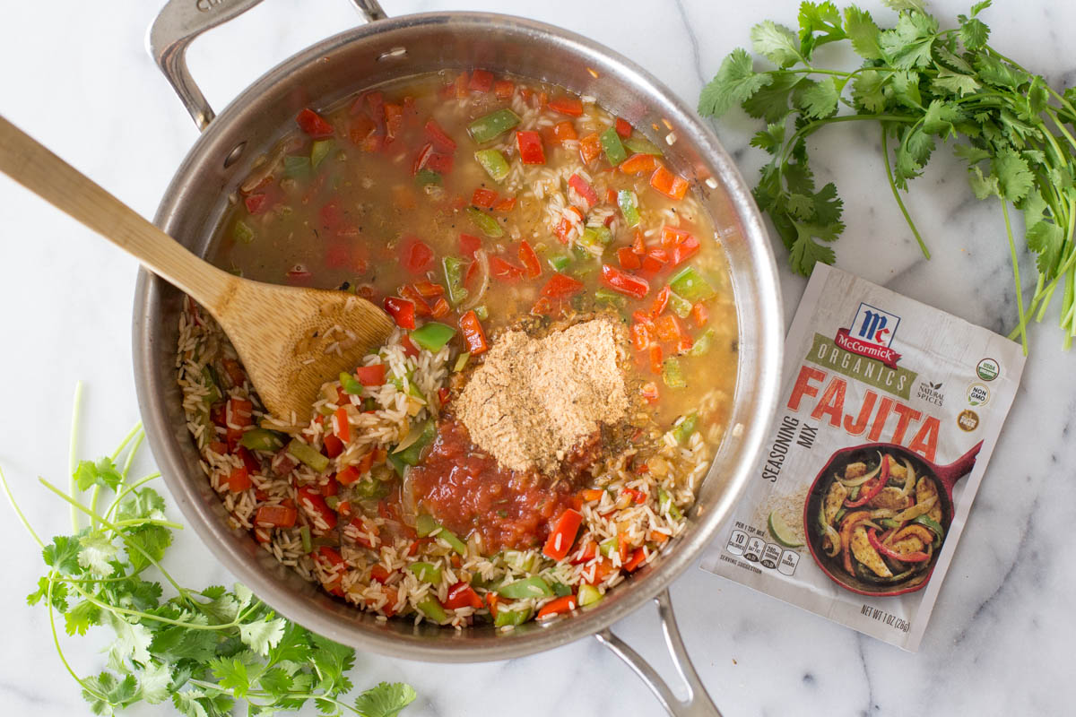 A skillet with the cooked onions and peppers, rice, chicken broth, salsa, the seasoning mix, and a wooden spoon, with a packet of the McCormick Organics Fajita Seasoning Mix next to it.