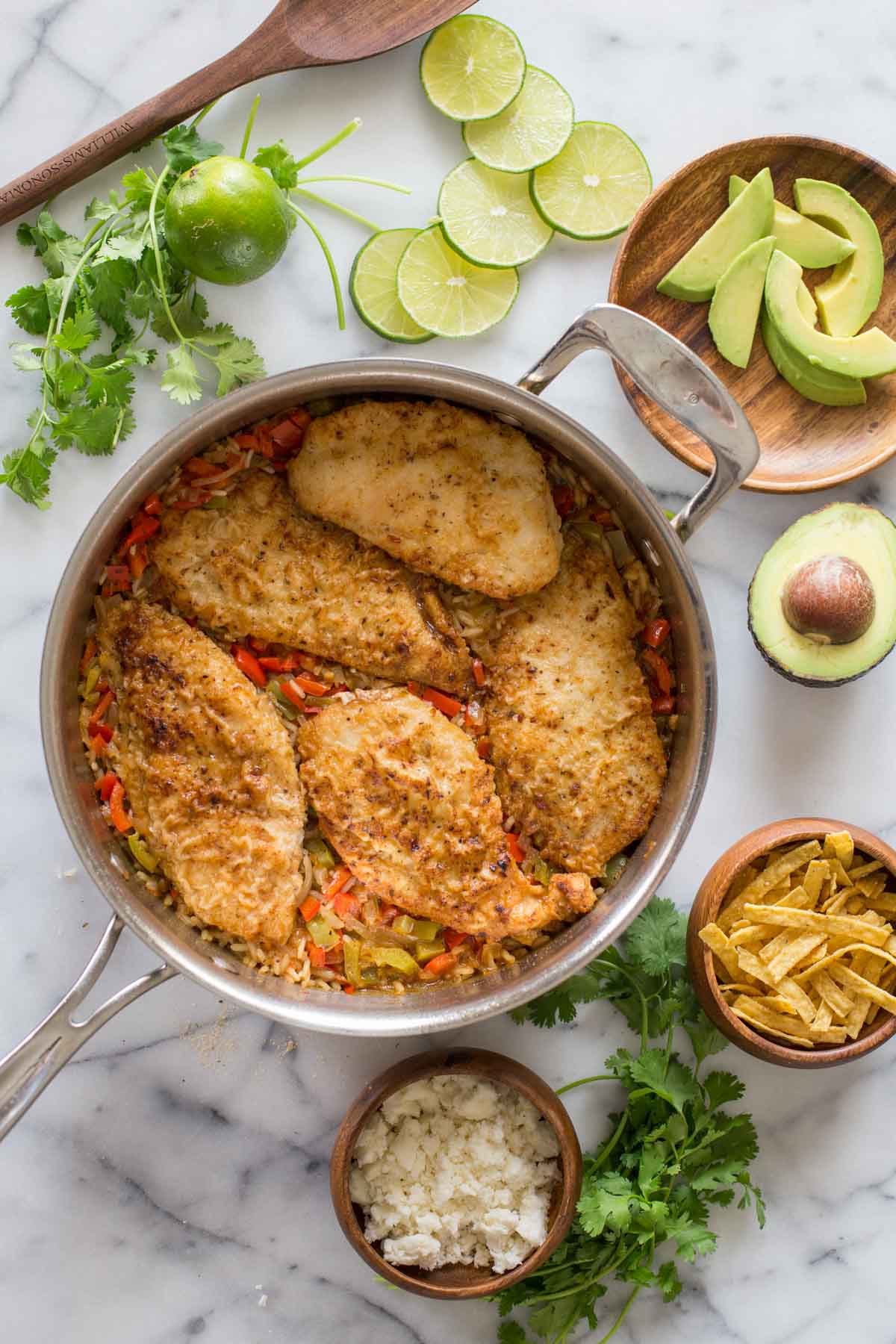 One Skillet Fajita Style Chicken and Rice, with a little bowl of crumbled queso fresco, fresh cilantro, a small bowl of tortilla strips, a half of an avocado, a plate of sliced avocado, some slices of lime, and a whole lime next to the skillet.