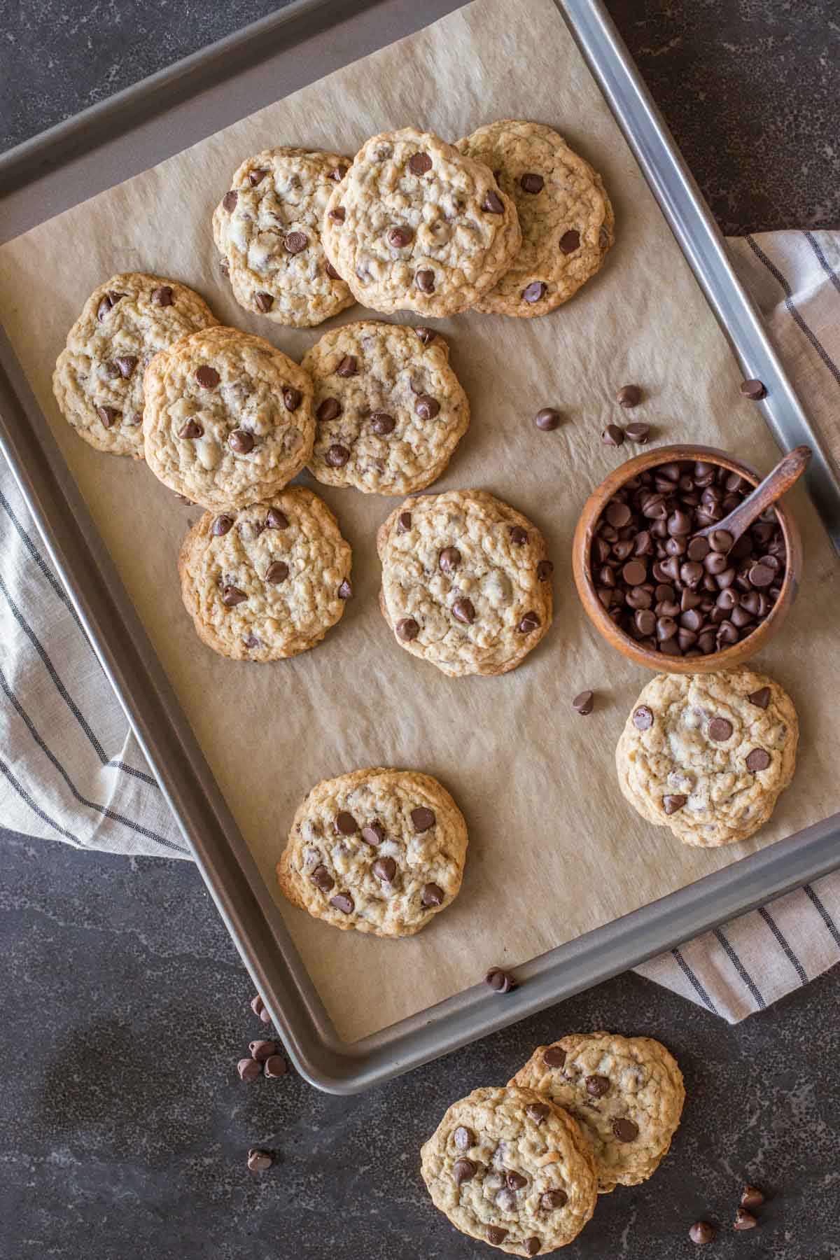 Bakery Style Oatmeal Chocolate Chip Cookies on a parchment paper lined baking sheet, along with a small wood bowl of chocolate chips, and two cookies sitting next to the baking sheet.