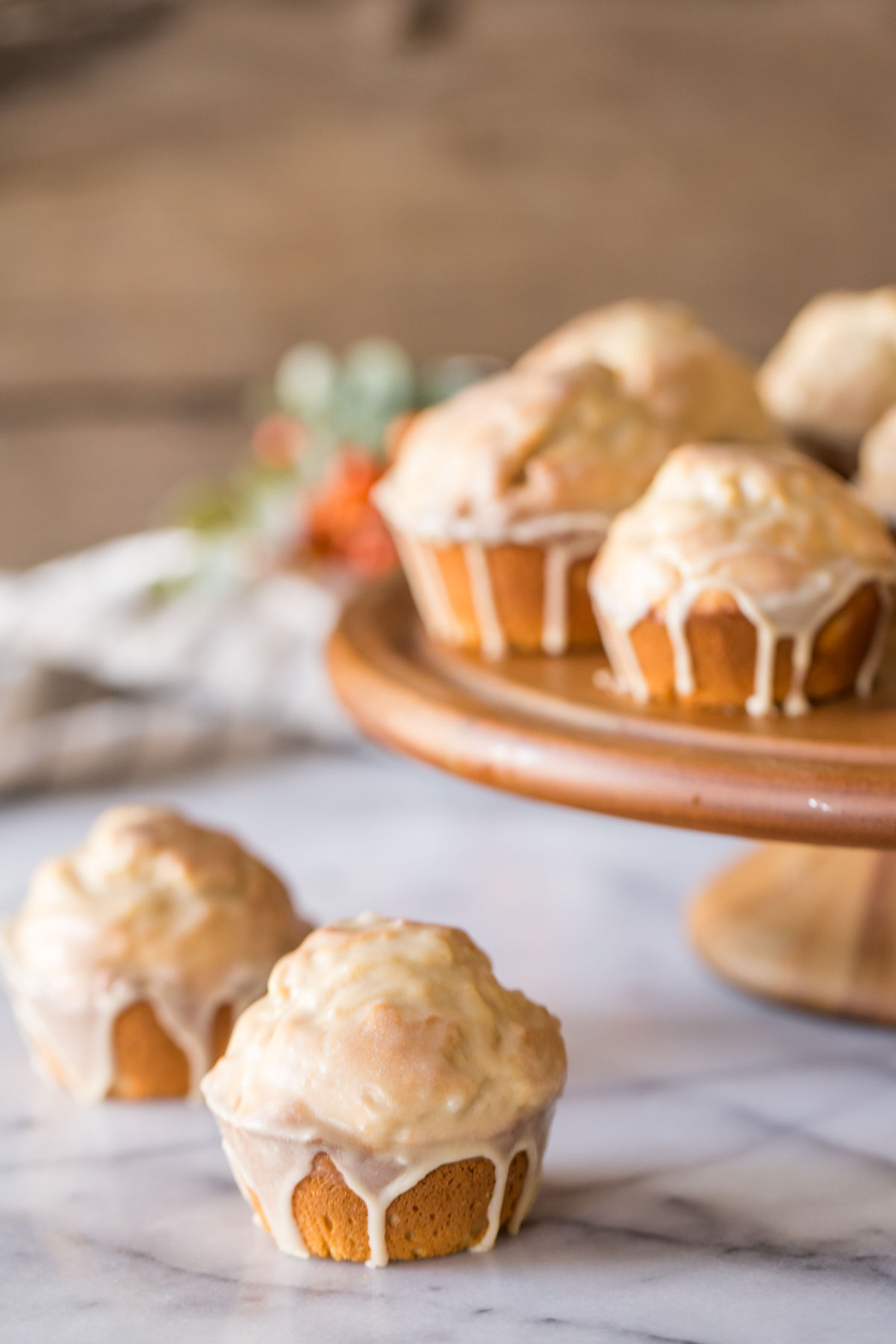 Two Maple Glazed Donut Muffins sitting next to a wood cake stand with more Maple Glazed Donut Muffins on it.
