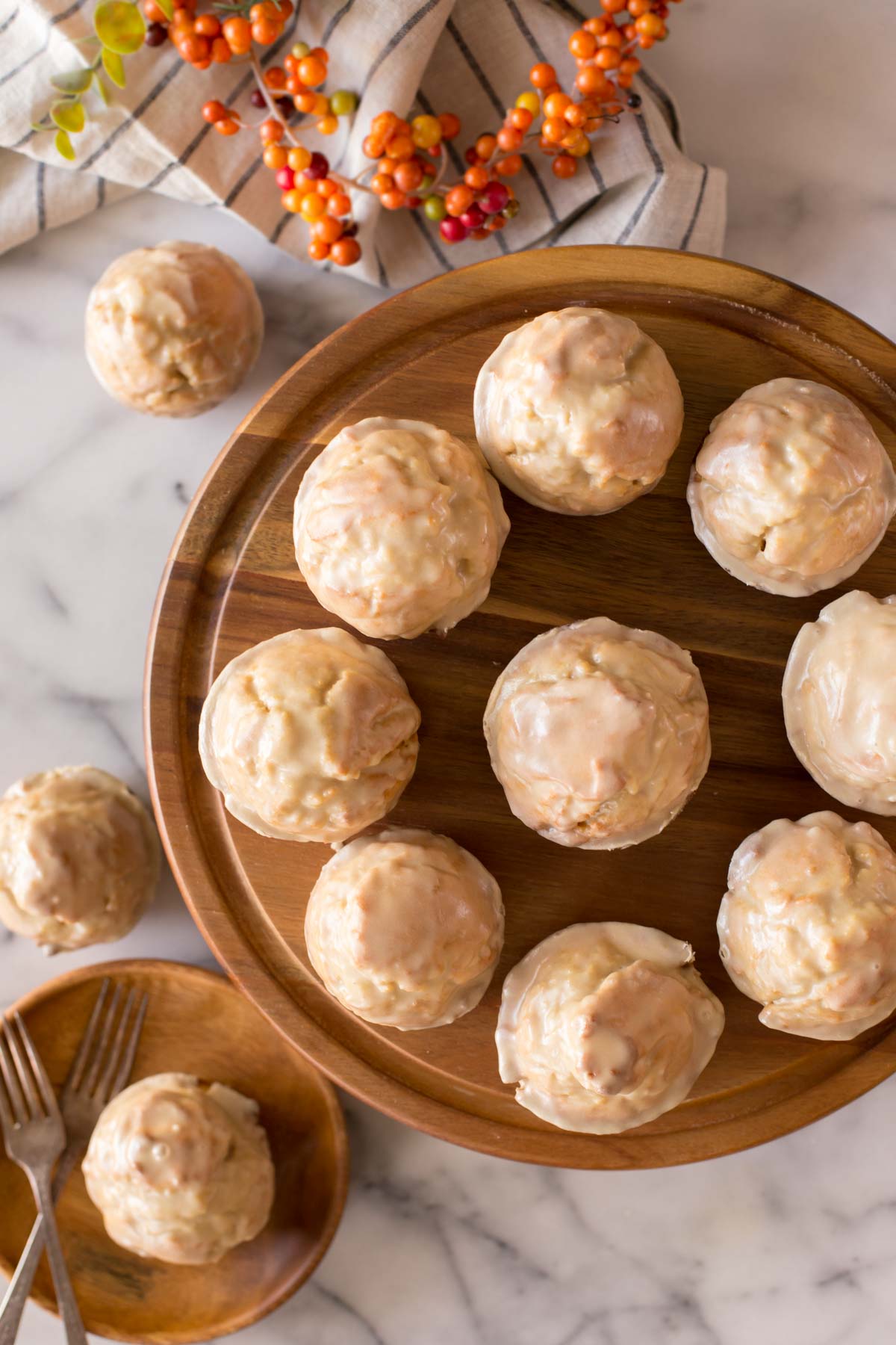 Maple Glazed Donut Muffins on a wood cake stand, with three Maple Glazed Donut Muffins sitting next to the cake stand, one of them on a wood plate with two forks.