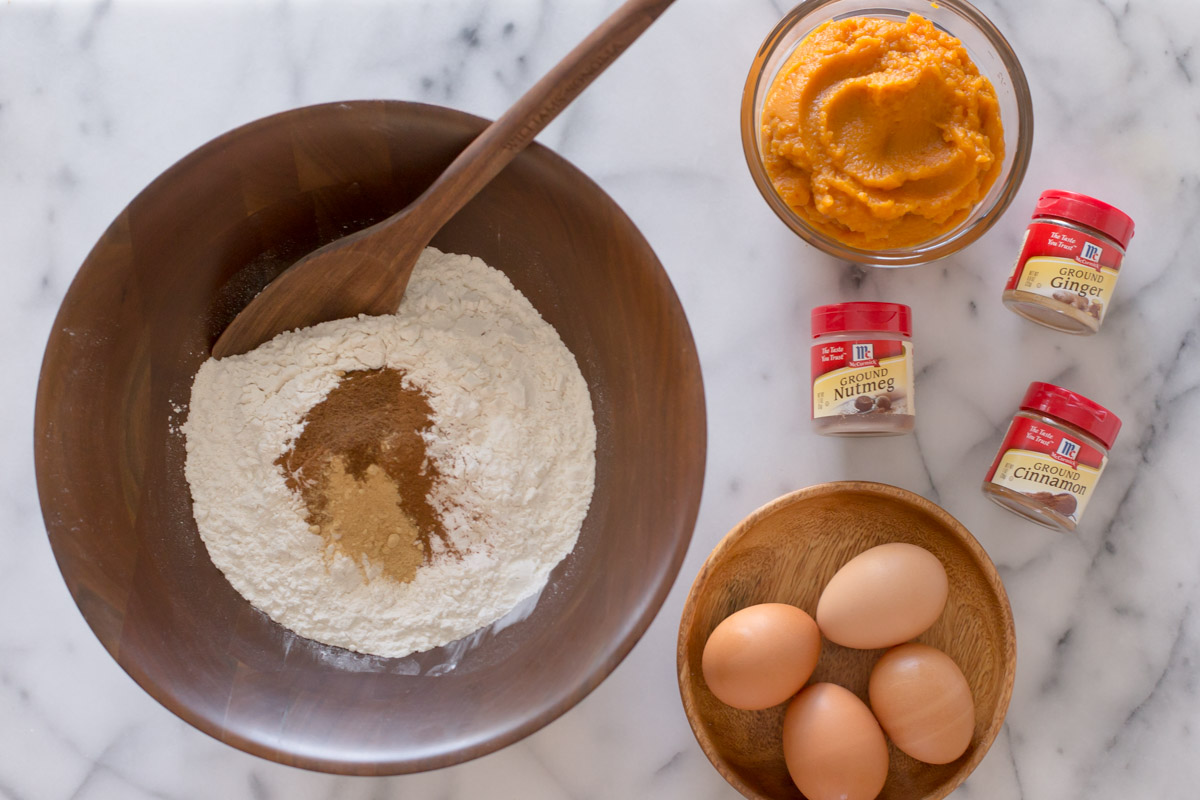A large wood bowl of the dry ingredients for the Pumpkin Spice Coffee Cake, sitting next to a glass bowl of pumpkin puree, three McCormick spice containers, and a wood plate with four eggs on it.