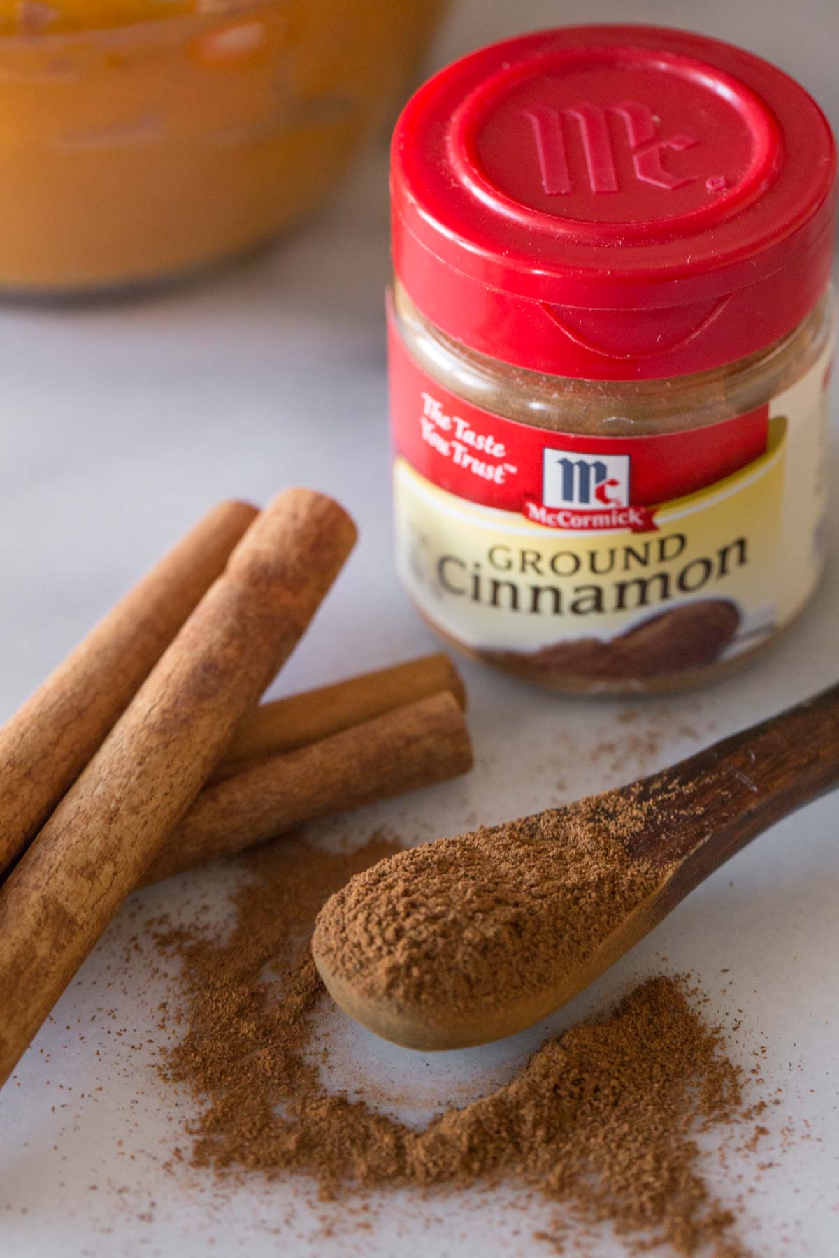 A McCormick spice container of Ground Cinnamon, with a few cinnamon sticks and a small wood spoon of ground cinnamon sitting in front of it.