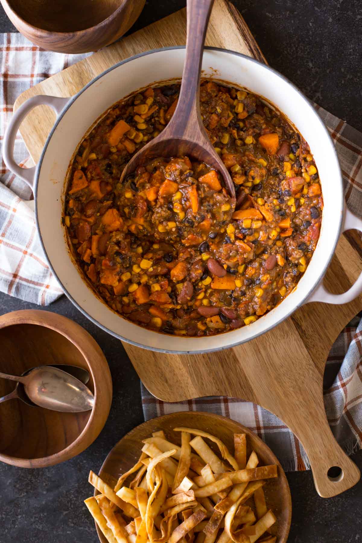 A large pot of Sweet Potato Chili with a wooden spoon in it, and a plate of Fried Tortilla Strips and wood bowls next to the pot.