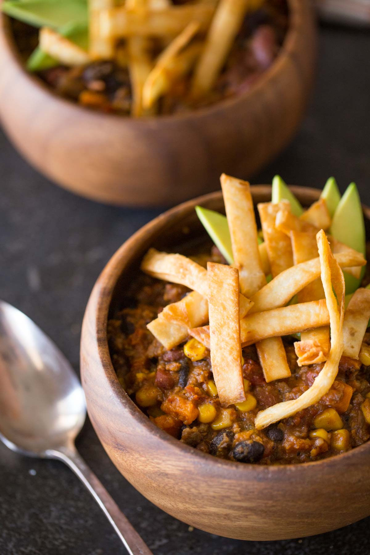 Two wood bowls of Sweet Potato Chili, topped with Fried Tortilla Strips and sliced avocado.