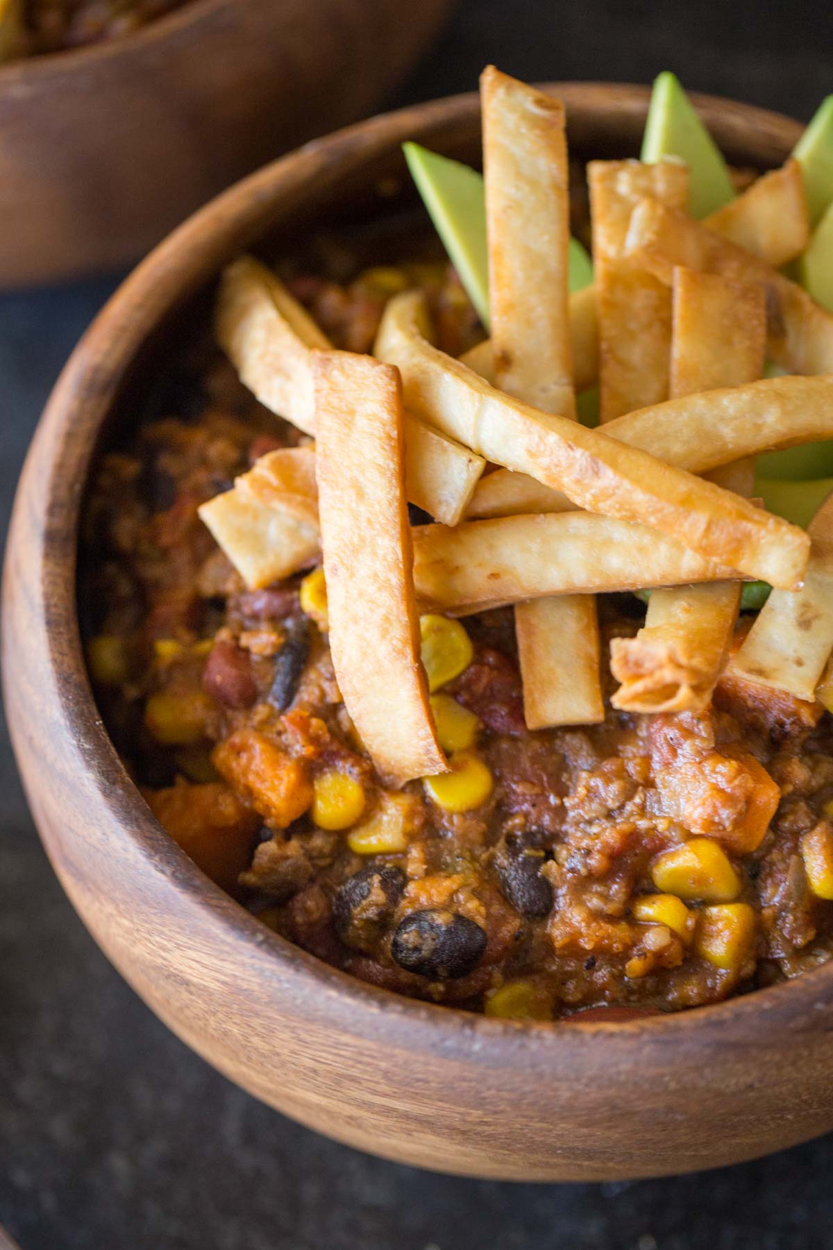 A wood bowl of Sweet Potato Chili, topped with Fried Tortilla Strips and sliced avocado.