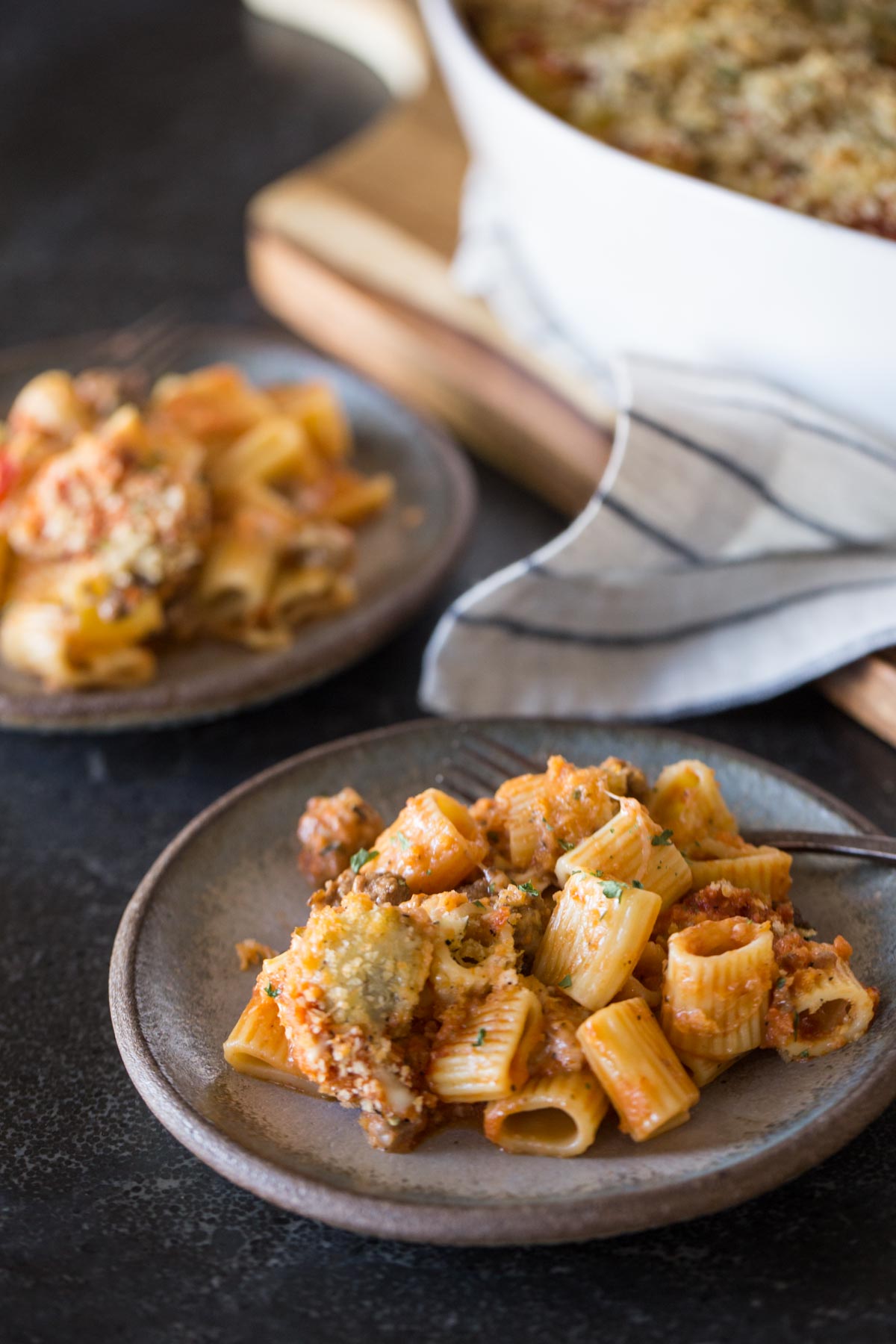 Two plates of Cheesy Tomato Alfredo Pasta Bake sitting next to the baking dish of Cheesy Tomato Alfredo Pasta Bake.