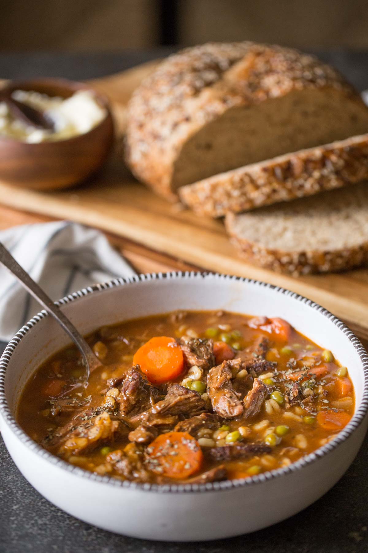Beef and Barley Vegetable Soup in a bowl with a spoon, sitting next to a cutting board with bread and a small bowl of soft butter. 