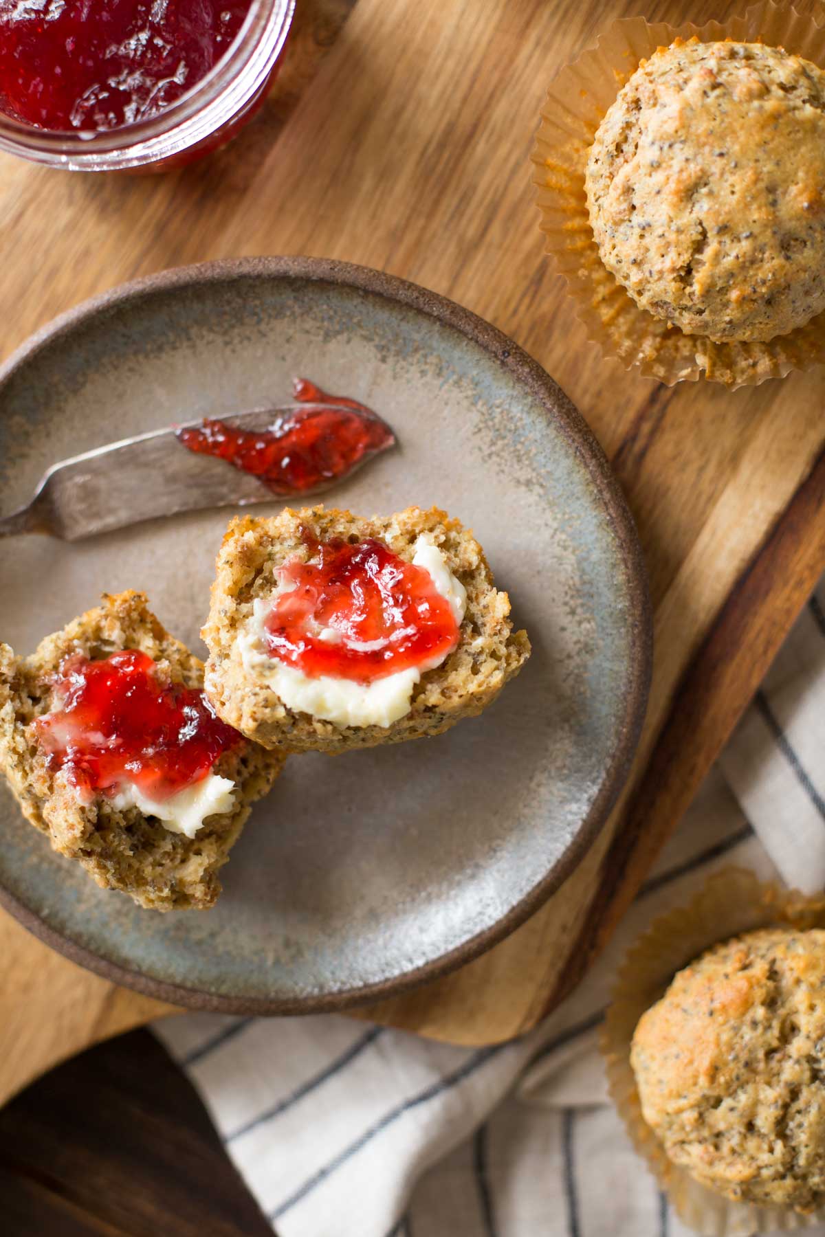 A Honey Bran Muffin cut in half with butter and jam on it, sitting on a plate with the jam knife, with two whole Honey Bran Muffins next to the plate along with the jar of jam.