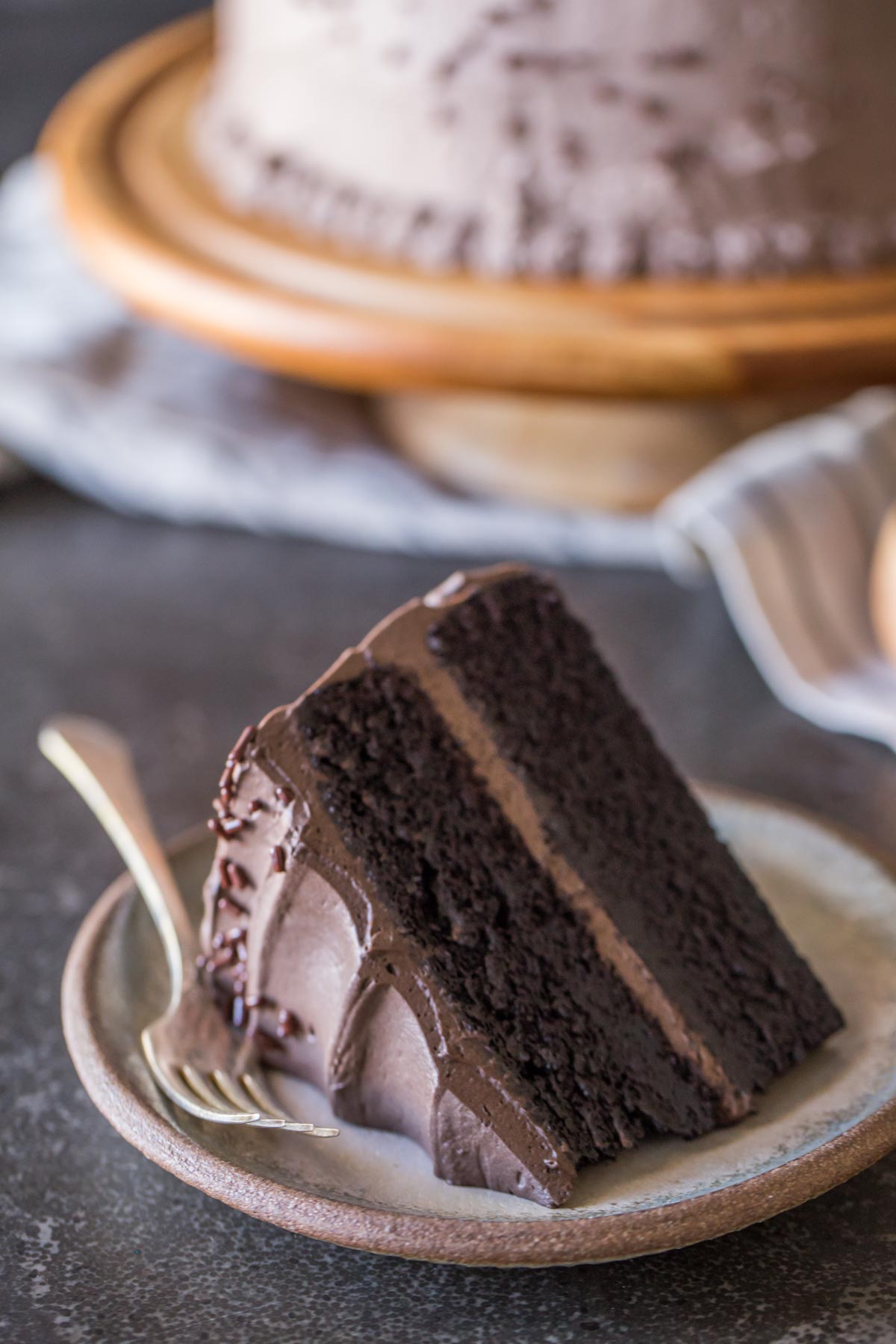 A slice of Dark Chocolate Cake With Whipped Cream Frosting on a plate with a fork, and the rest of the cake on a cake stand in the background.