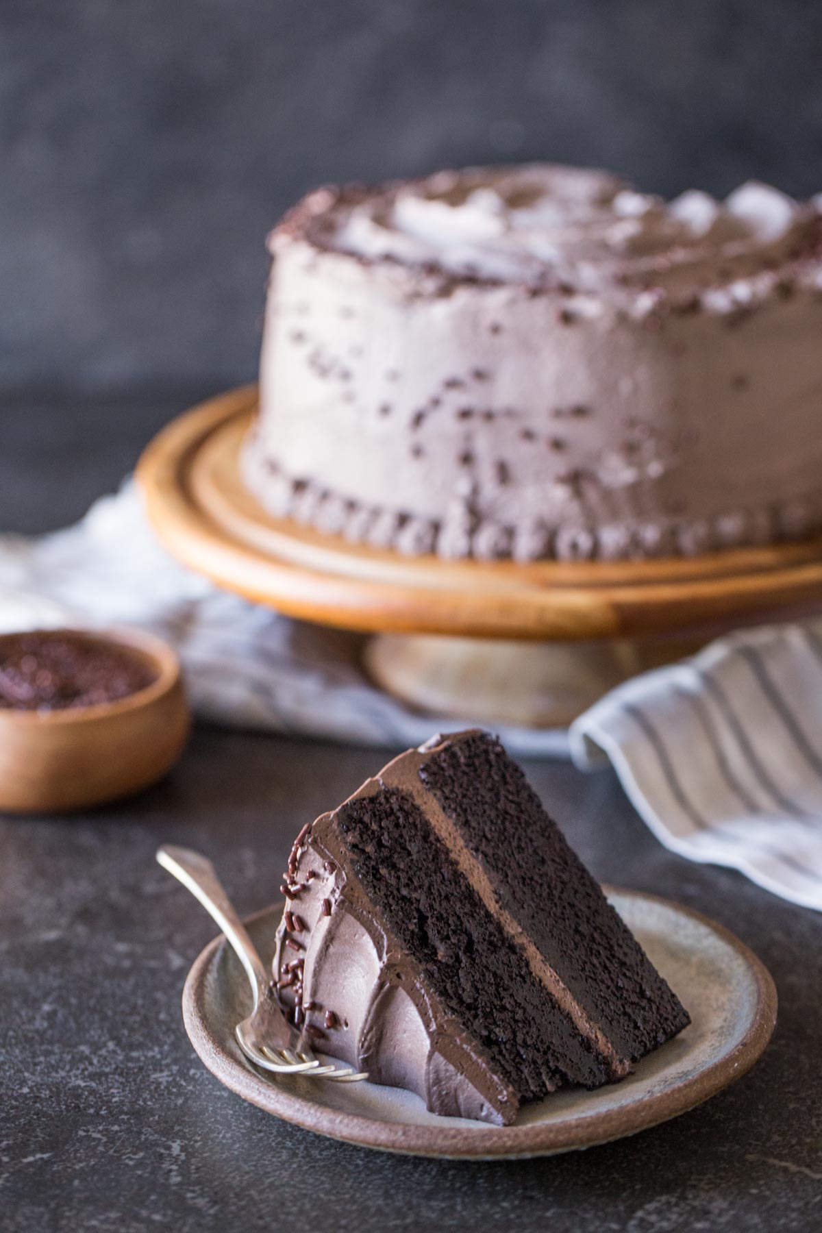 A slice of Dark Chocolate Cake With Whipped Cream Frosting on a plate with a fork, and the rest of the cake on a cake stand in the background along with a small wood bowl of chocolate sprinkles.