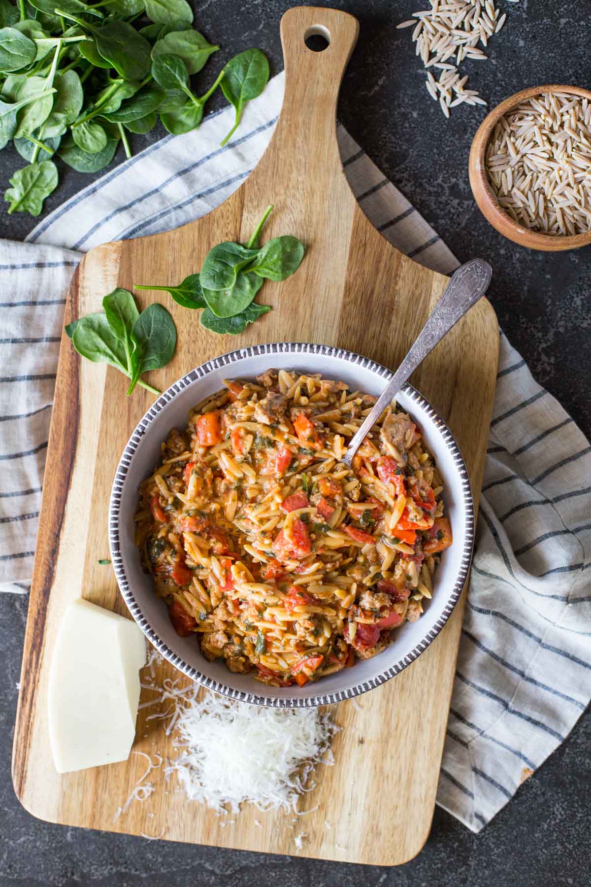 One Pot Creamy Tomato Orzo in a bowl, sitting on a board with some freshly grated parmesan cheese and some baby spinach, with a small wood bowl of uncooked whole wheat orzo next to the board.