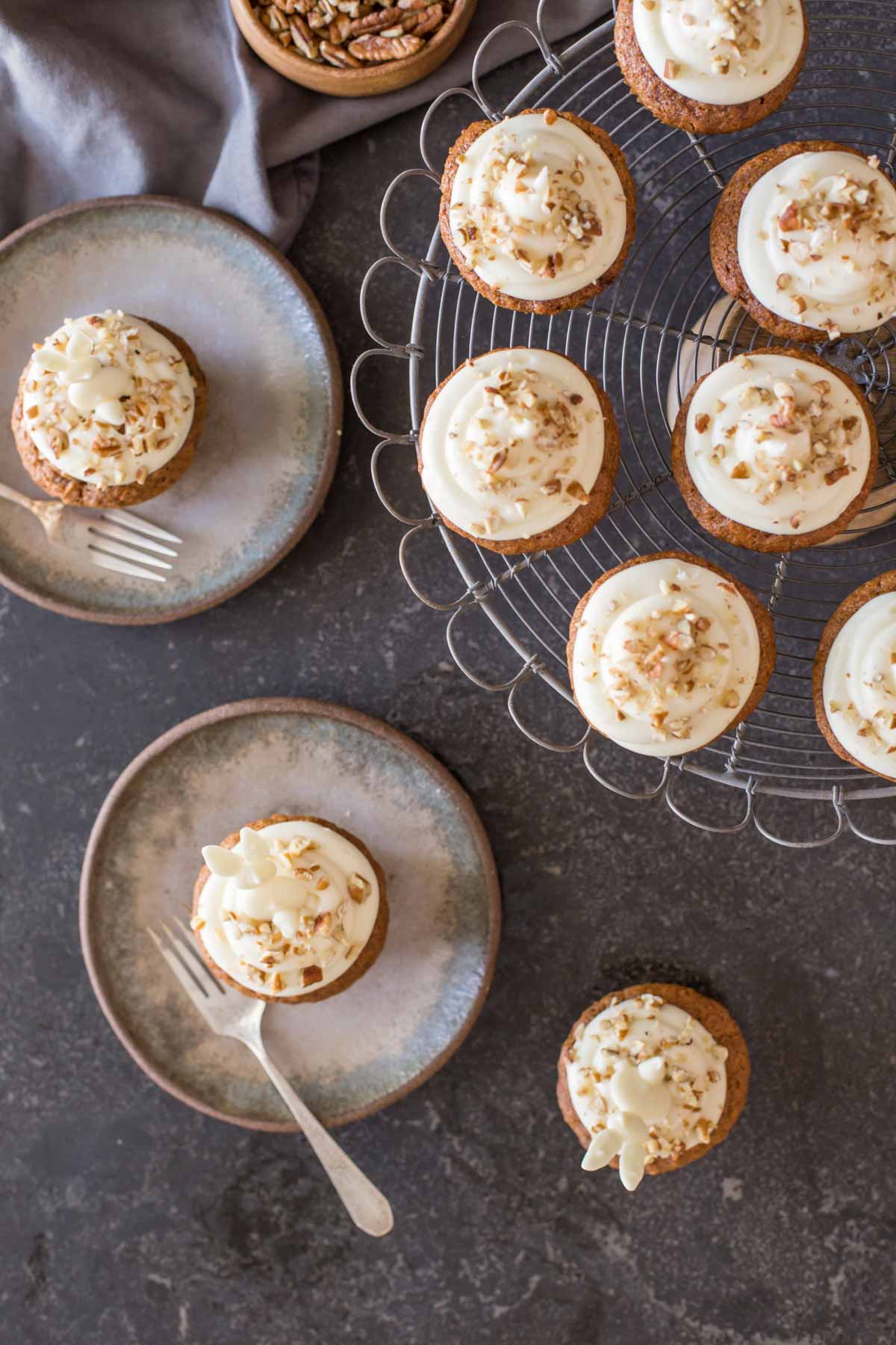 Three Carrot Cake Cupcakes with white chocolate bunnies on top, two of them sitting on plates with forks, all sitting next to a cake stand with more Carrot Cake Cupcakes.