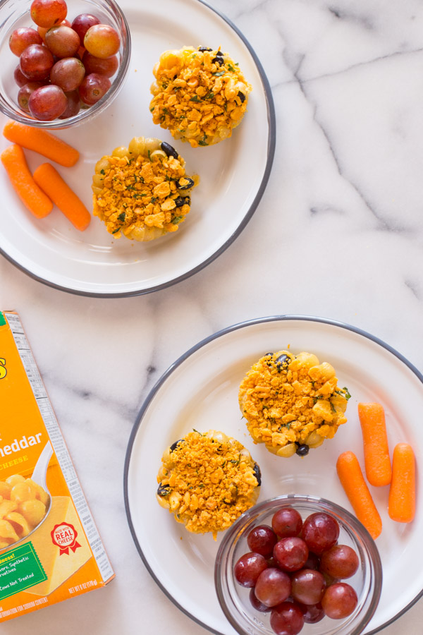 Two plates with two Southwestern Mac and Cheese cups on each plate, along with a cup of grapes and some carrot sticks.