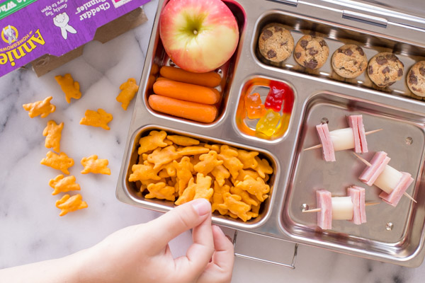 A lunch box with an apple, carrot sticks, Annie's Cheddar Bunnies, Annie's Cookie Bites, some gummy snacks, and ham and cheese on toothpicks.