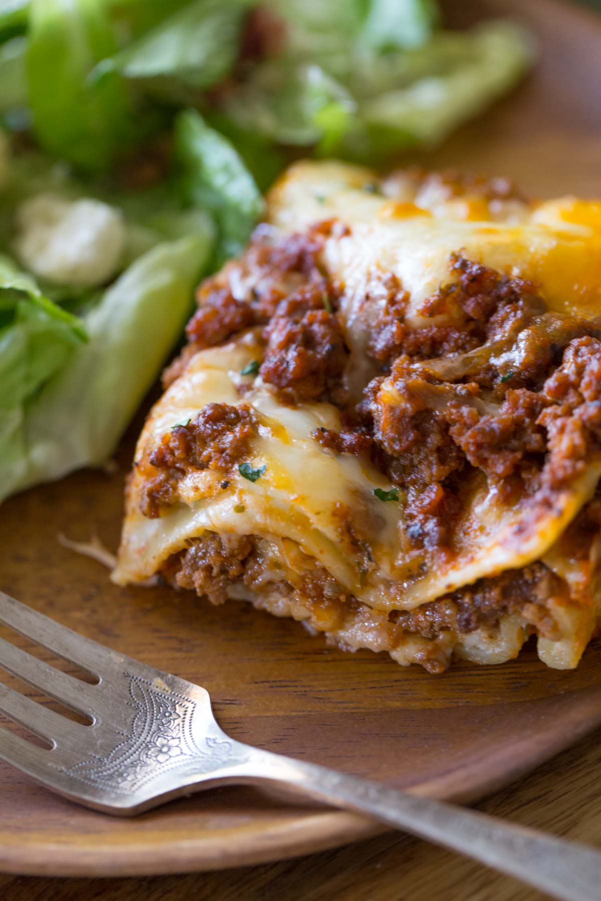A piece of Simple Homemade Lasagna on a wood plate with a fork and salad.