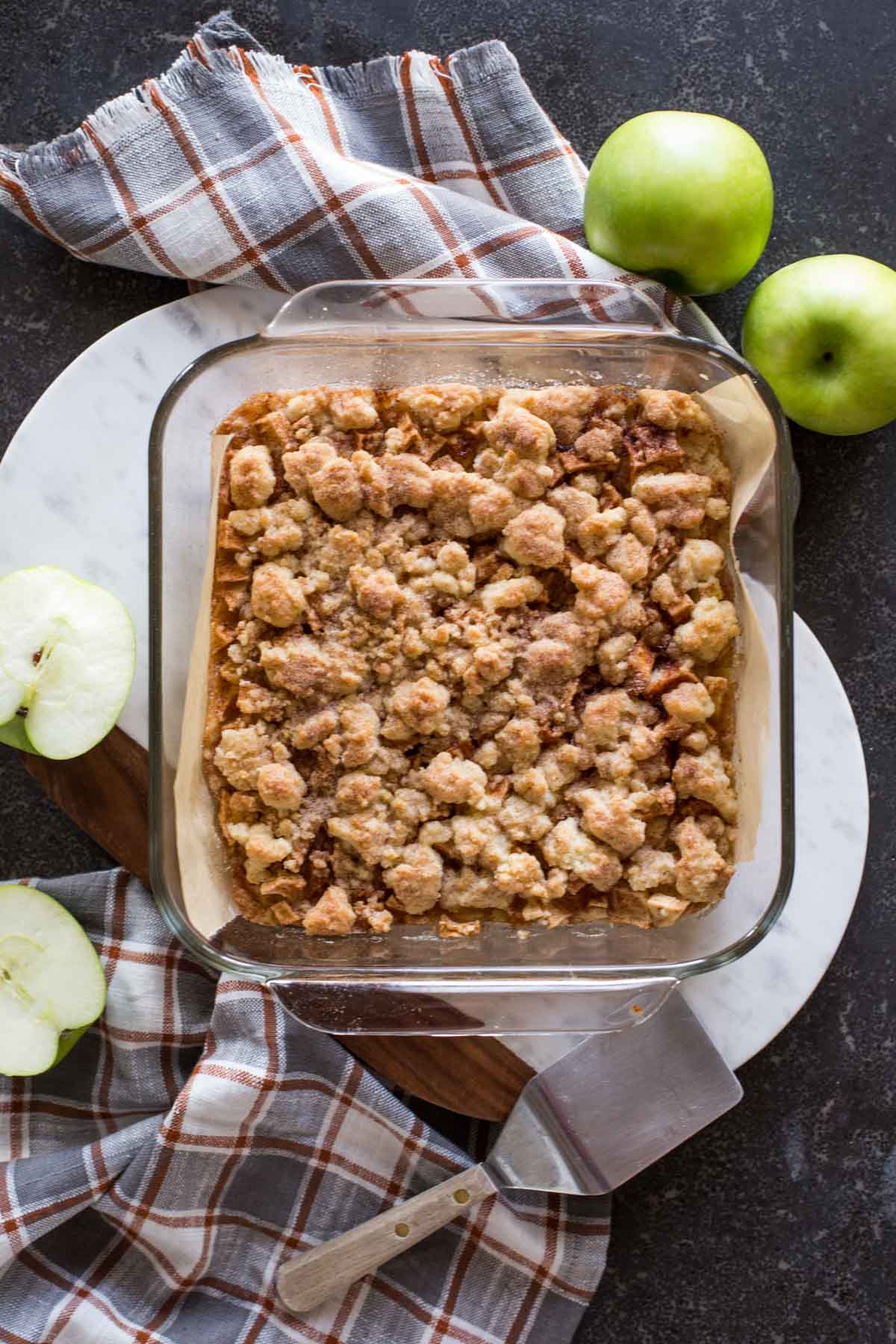 Apple Pie Crumb Bars in a square baking dish, with whole and halved apples next to it, as well as a serving spatula.
