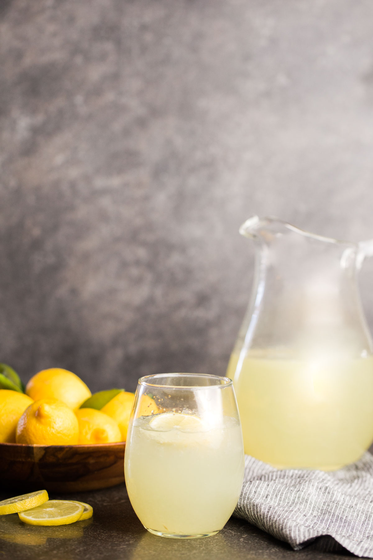 Lemonade in glass pitcher with bowl of lemons and glass of lemonade.