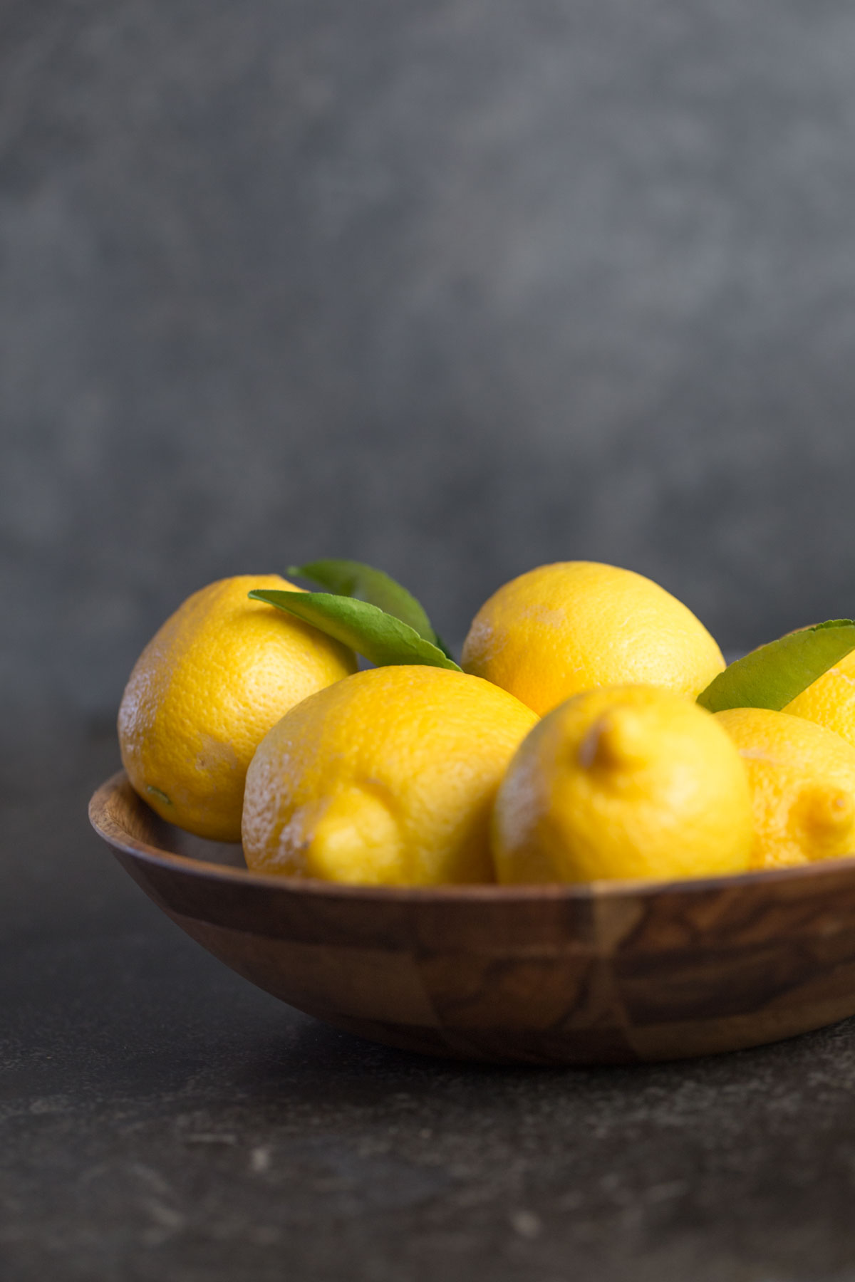 Wooden bowl of lemons with leaves attached from the front view with a dark grey background.