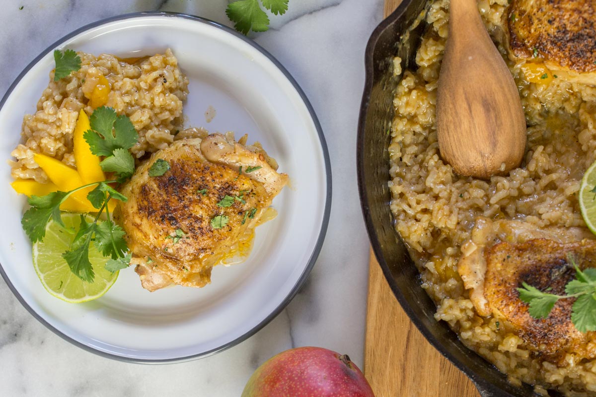 A serving of Chili Lime Mango Chicken and Rice on a plate, garnished with fresh cilantro, mango and lime slices, sitting next to the cast iron skillet of Chile Lime Mango Chicken and Rice. 