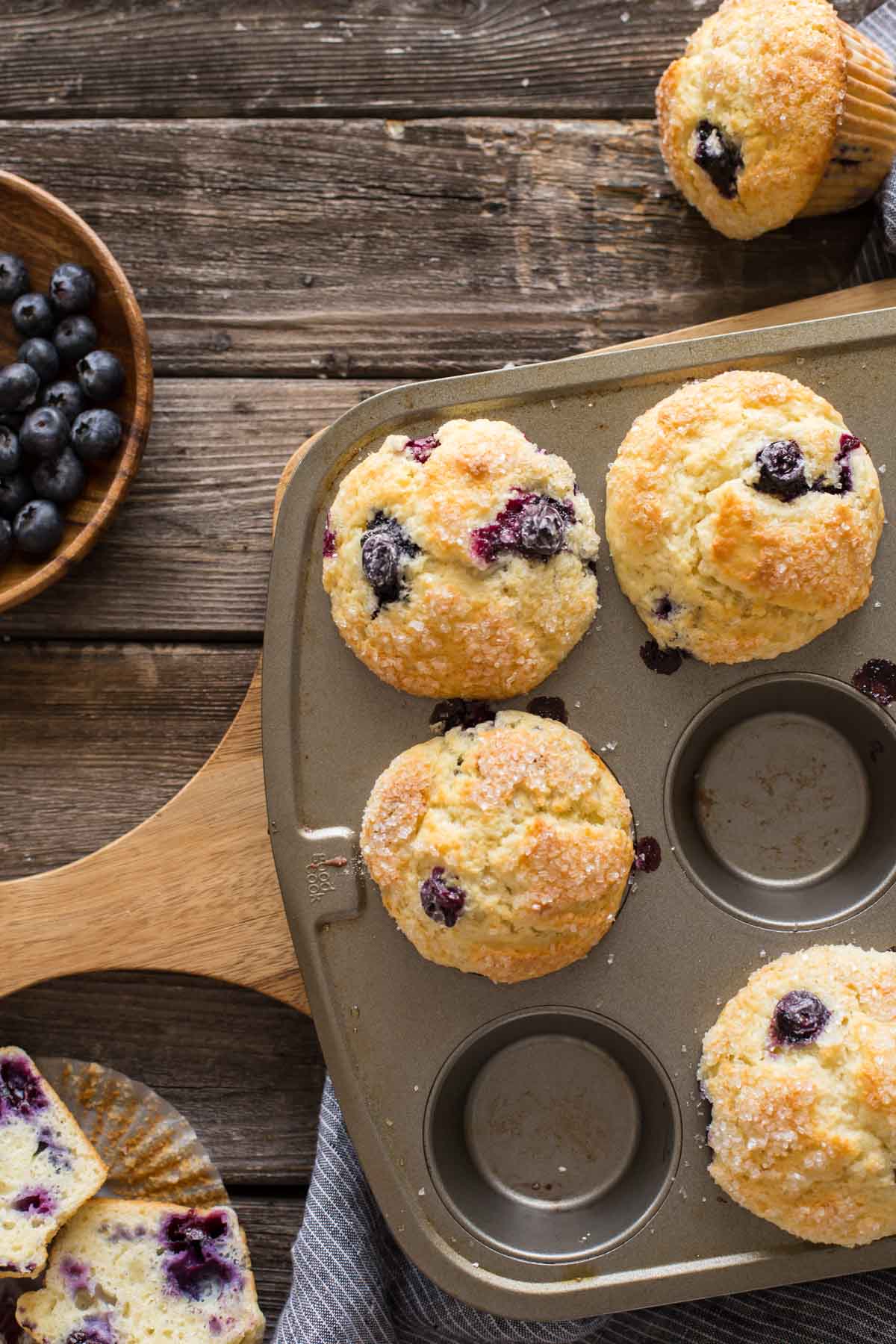 Best Ever Buttermilk Blueberry Muffins in a muffin pan on a wooden backboard with a bowl of fresh blueberries.