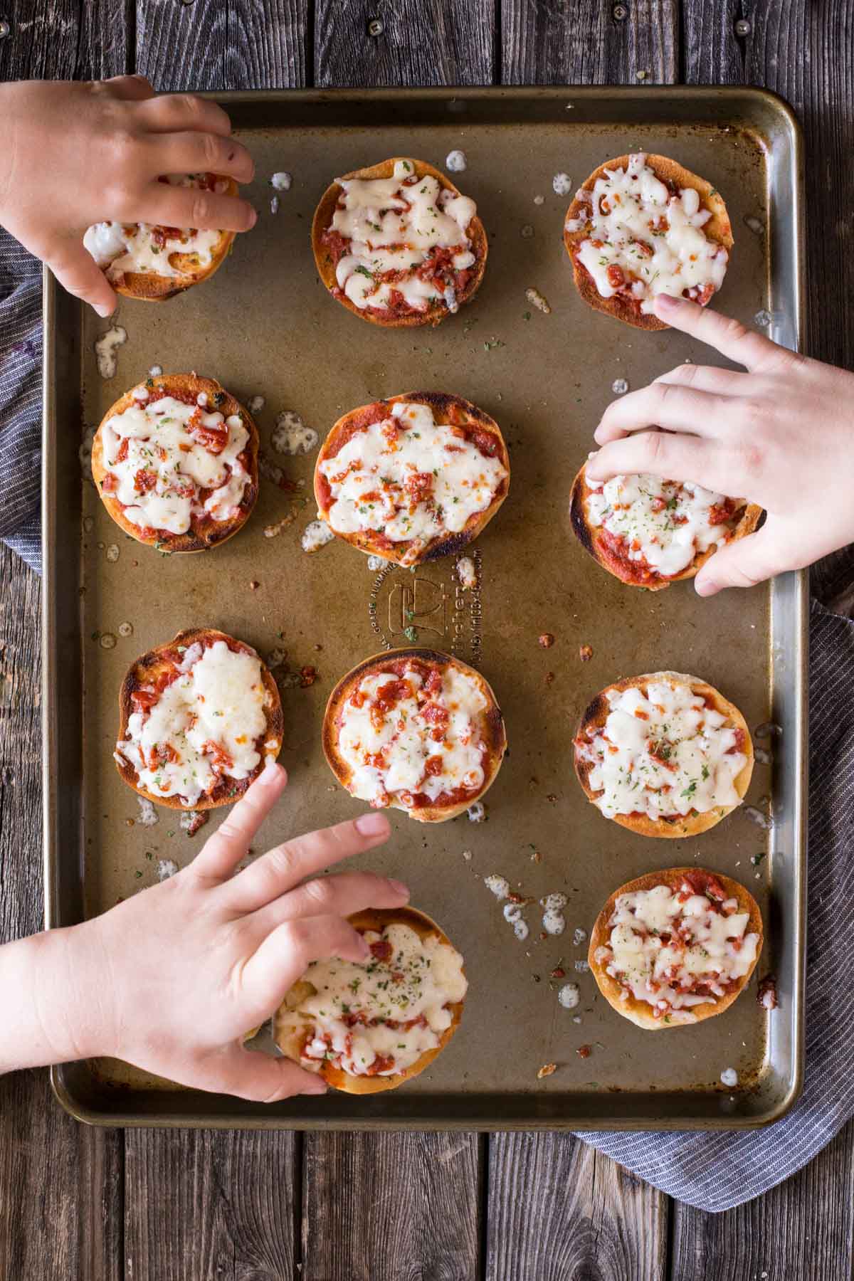 Top down view of Homemade Mini Bagel Pizzas on a baking sheet with hands reaching in.