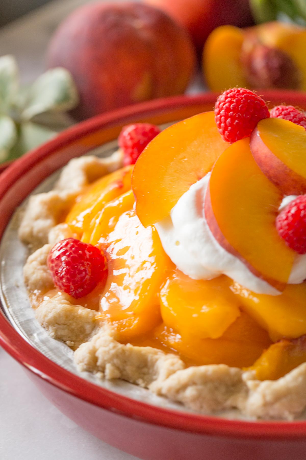 A close up shot of Fresh peach pie with homemade whipped cream in a red pie dish on a white marble background.