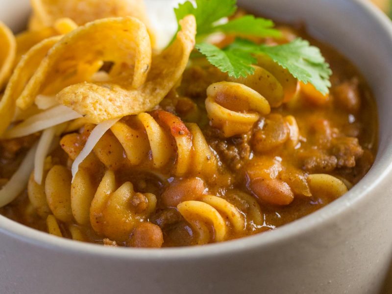 A close up shot of a grey bowl filled with Beef Chili Queso with pasta, Fritos, source cream, and cilantro.
