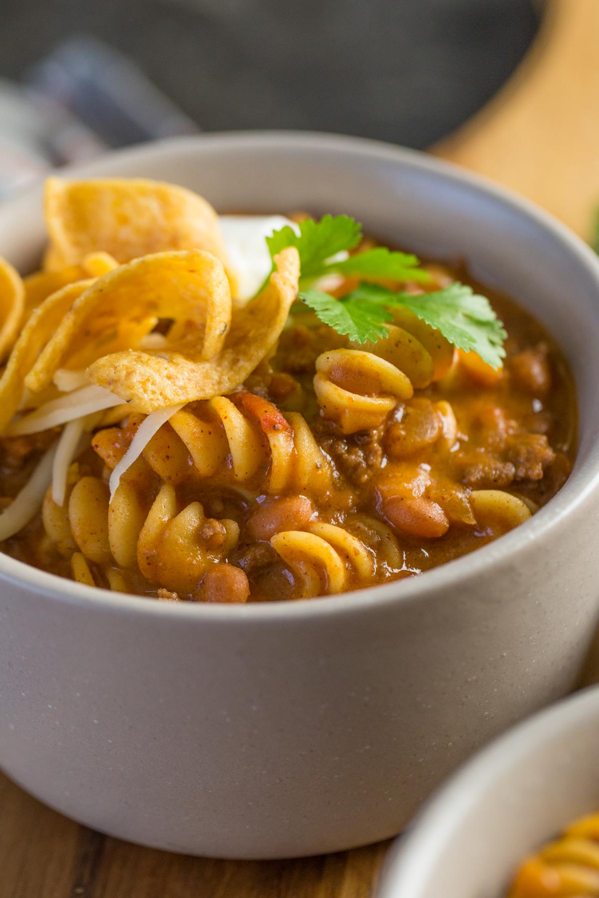 A close up shot of a grey bowl filled with Beef Chili Queso with pasta, Fritos, source cream, and cilantro.