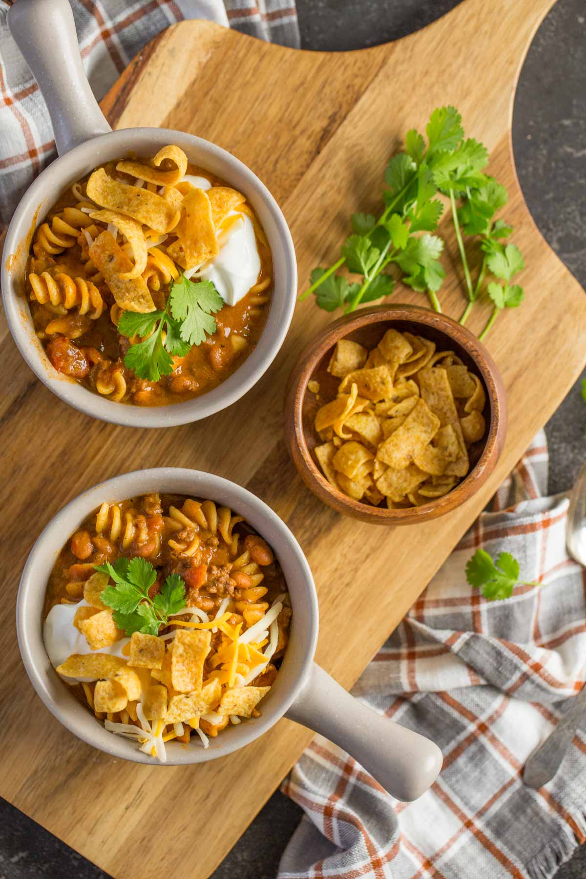 A top down view of two bowls Beef Queso Chili topped with sour cream, Fritos and Cilantro on a wooden board. 