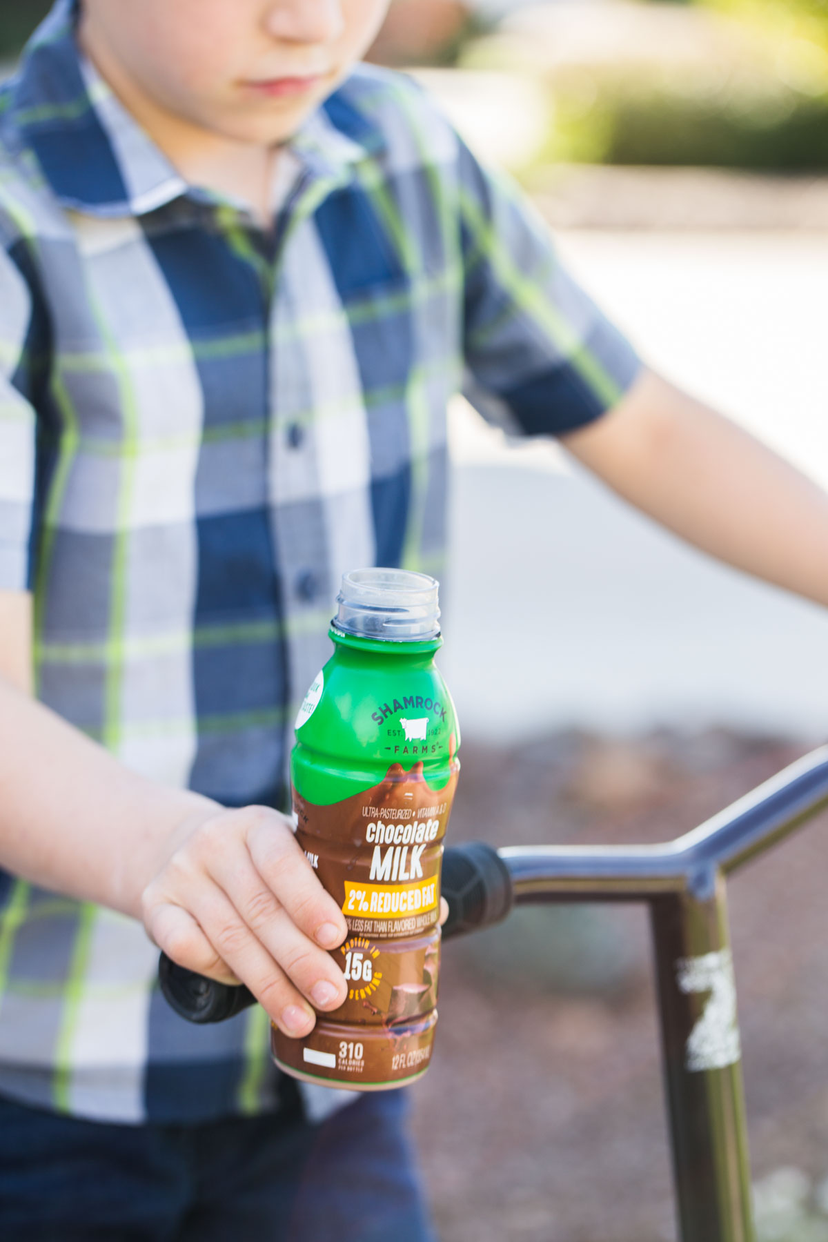 Boy riding a scooter with a single serve chocolate milk in one hand.