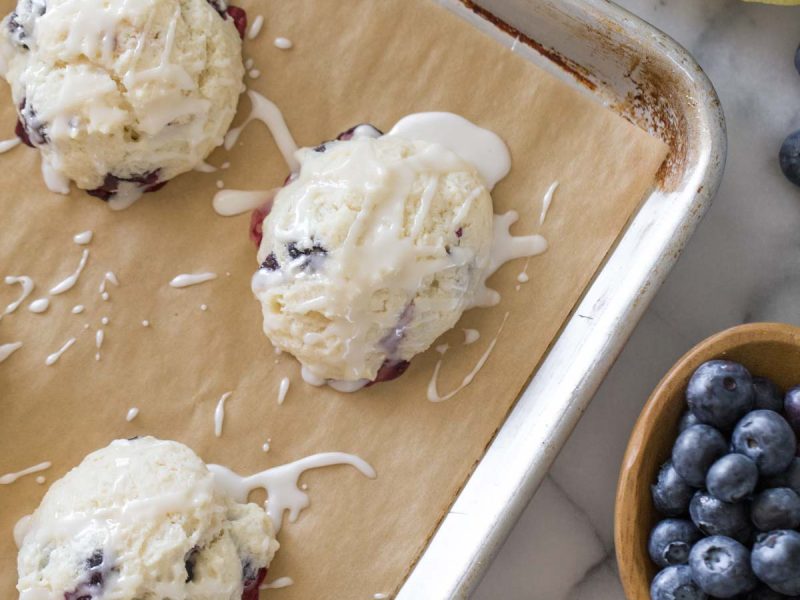 Glazed Lemon Blueberry Muffin Tops on a baking sheet with a grey background.