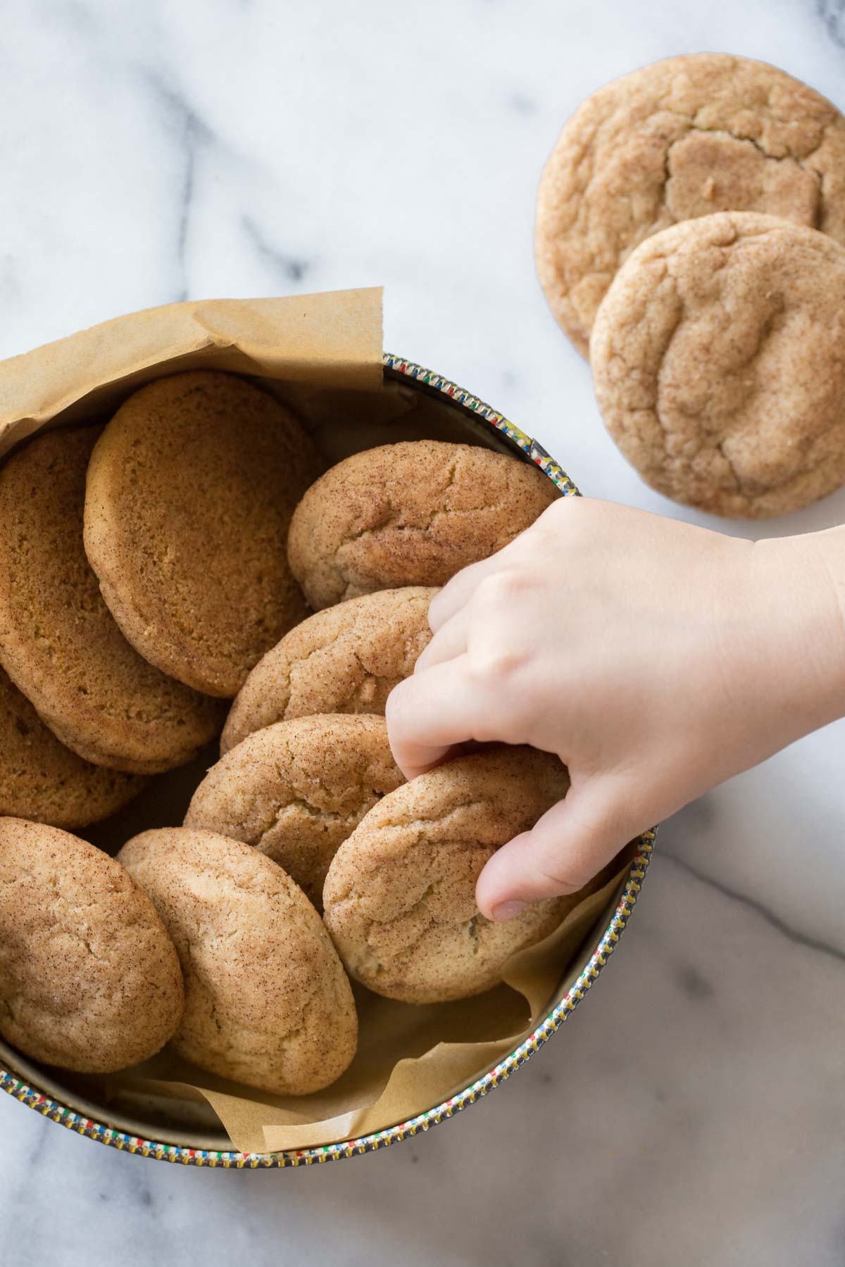 A child's hand reaching out to take a Super Soft Snickerdoodle cookie in a cookie tin on a white background.