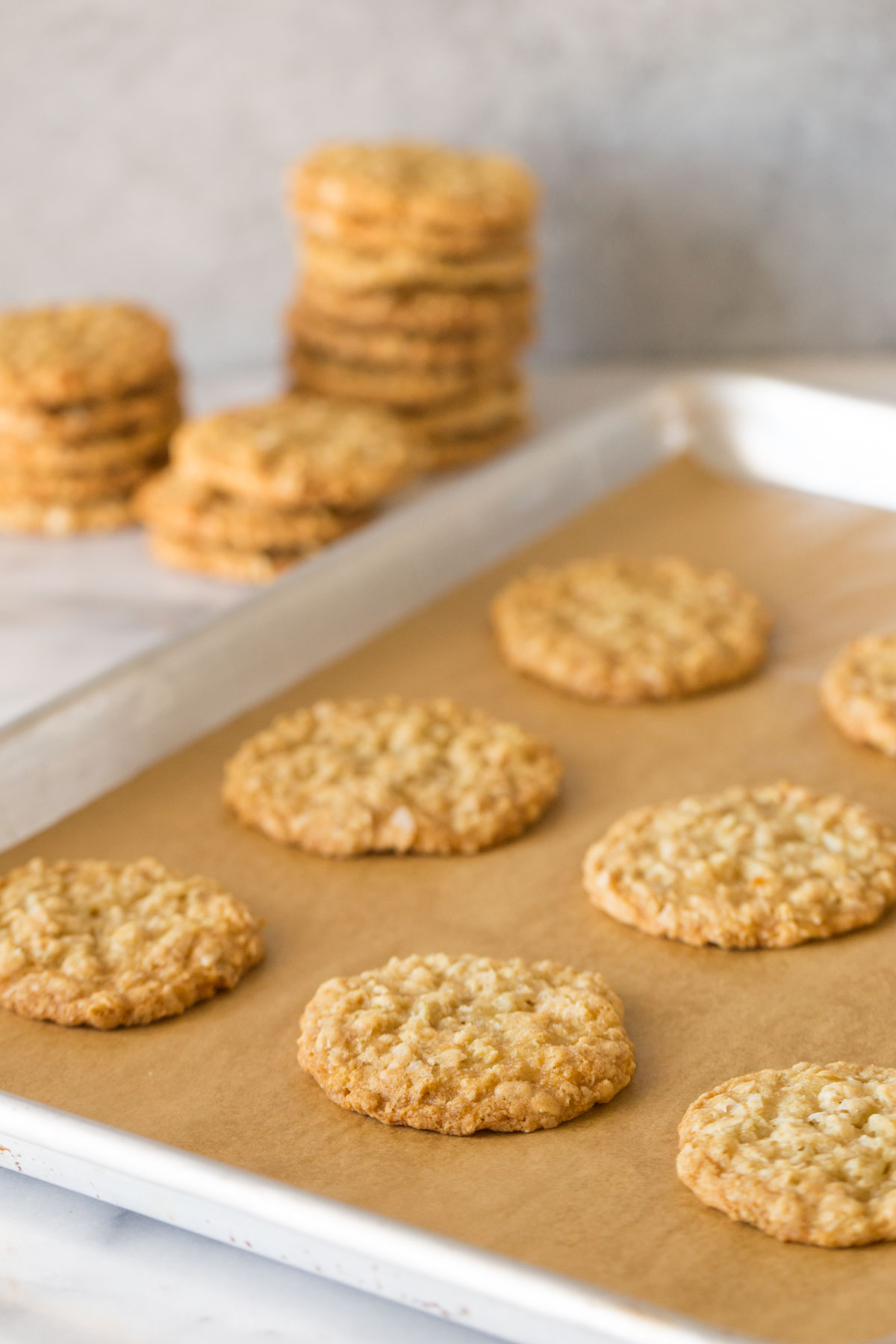 Buttery Coconut Oatmeal Cookies on a parchment paper lined baking sheet, with stacks of Buttery Coconut Oatmeal Cookies in the background.