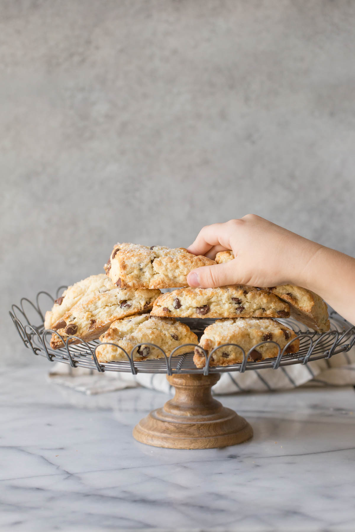 A stack of Chocolate Chip Scones on a cake stand with a hand reaching for the scone on top.